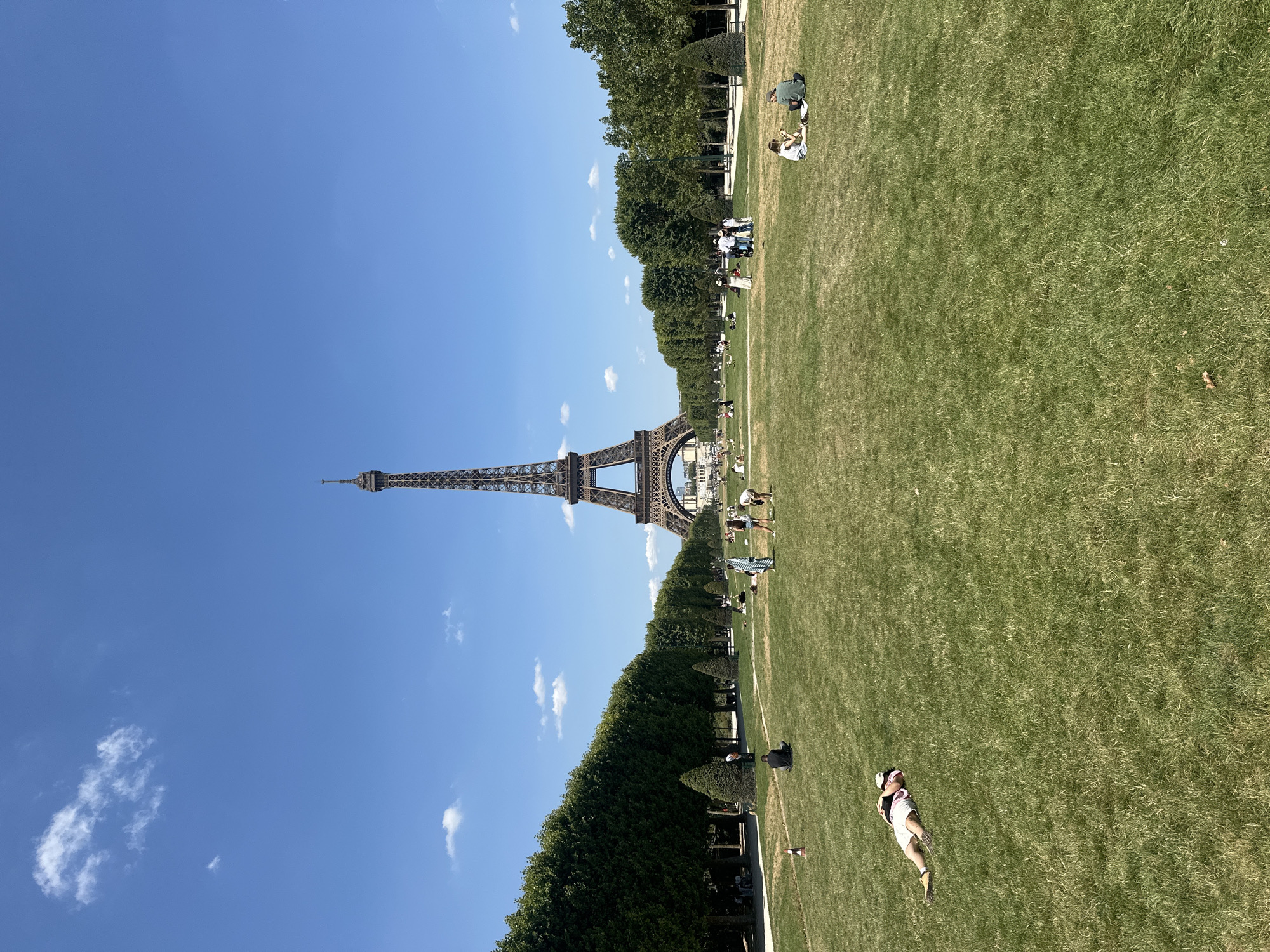 The image depicts the Eiffel Tower in Paris, France, on a clear, sunny day. The tower is prominently positioned in the background, dominating the skyline. In the foreground, a vast expanse of green grass is dotted with people engaged in various leisurely activities. Some individuals are seen relaxing on the lawn, while others are gathered in small groups, perhaps picnicking or socializing. A man is lying on his back on the grass, wearing shorts and a cap, appearing to rest or sunbathe. To the left and right of the Eiffel Tower, manicured rows of trees line pathways, creating a symmetrical and orderly park setting. The sky is a vibrant blue with scattered white clouds.