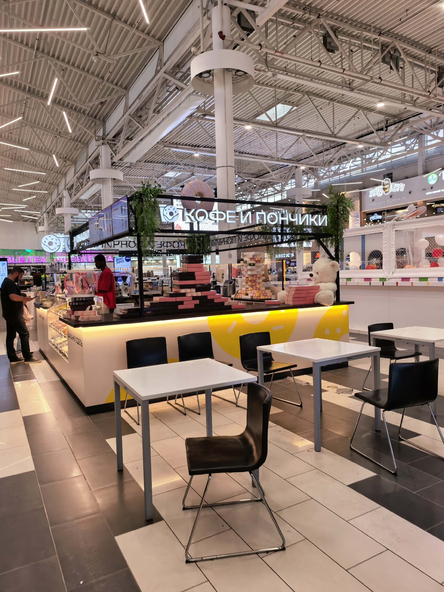 A bright, wide-angle interior shot captures a donut and coffee shop in Ufa, Russia, likely within a modern shopping mall given the high ceilings with exposed industrial beams and extensive fluorescent lighting. The shop's counter, emblazoned with the Russian text "КОФЕ И ПОНЧИКИ" (Coffee and Donuts), is brightly lit with a yellow underglow, highlighting stacks of pink donut boxes. A large, plush white teddy bear sits prominently on the counter next to a display of donuts and pastries. Two employees are visible: one in a red shirt, interacting with the display, and another in a black shirt, standing by the entrance, looking at a phone. Several white tables with black chairs are arranged in the foreground and mid-ground, some empty and some with partial views of customers or their belongings. The floor is a patterned mix of light and dark grey tiles. In the background, a children's play area with white netting and colorful accents is visible, suggesting the shop is in a family-oriented section of the mall. The overall impression is of a clean, well-lit, and contemporary food establishment.