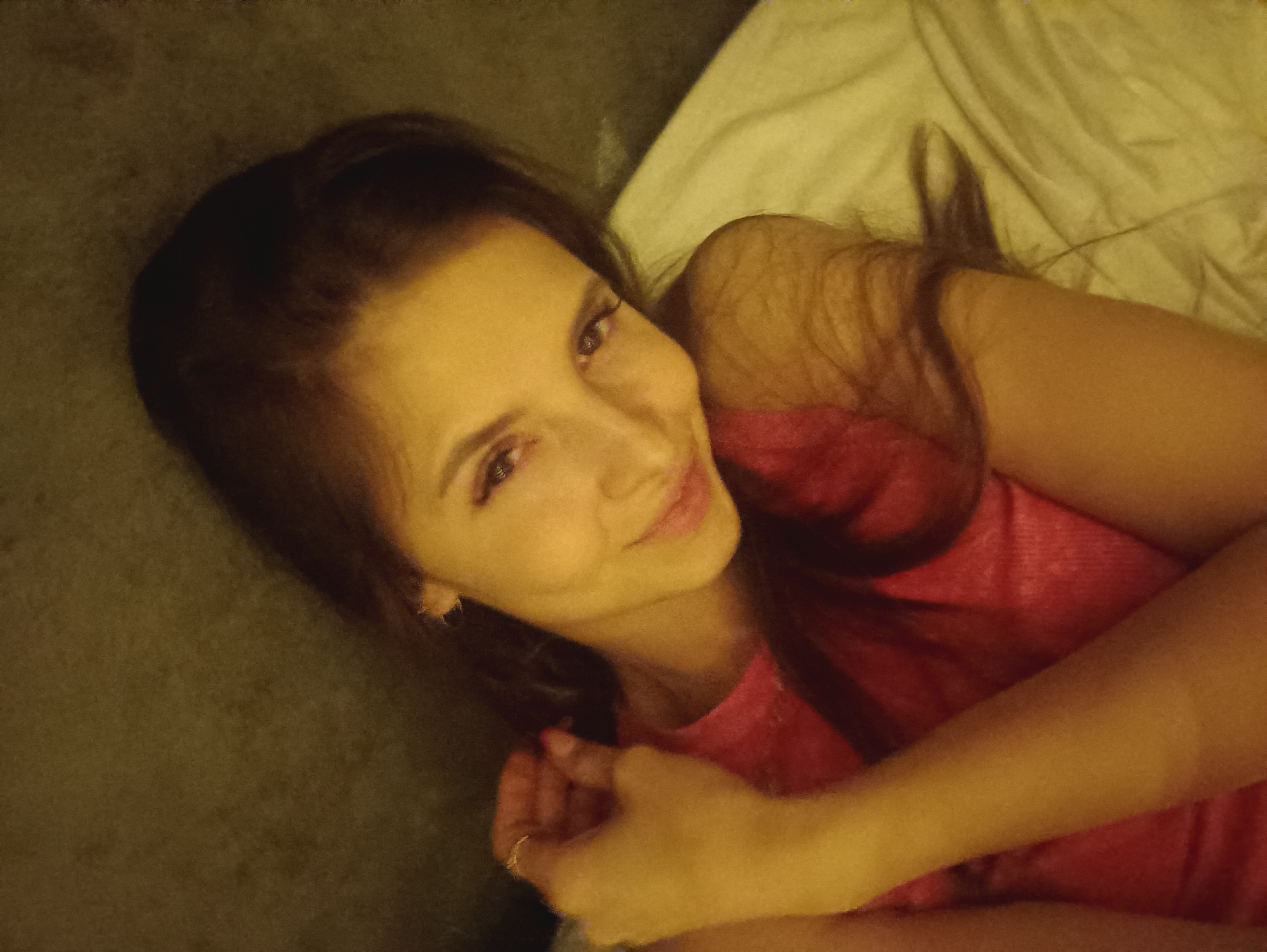A young woman is lying on a light-colored bed, wearing a pink top. She has long, dark hair and is smiling at the camera. She is clasping her hands together, with a ring visible on one finger. The background is indistinct, with a dark green-gray area and the white bedclothes. The location is not identifiable as Chaikovsky, Russia from the image.