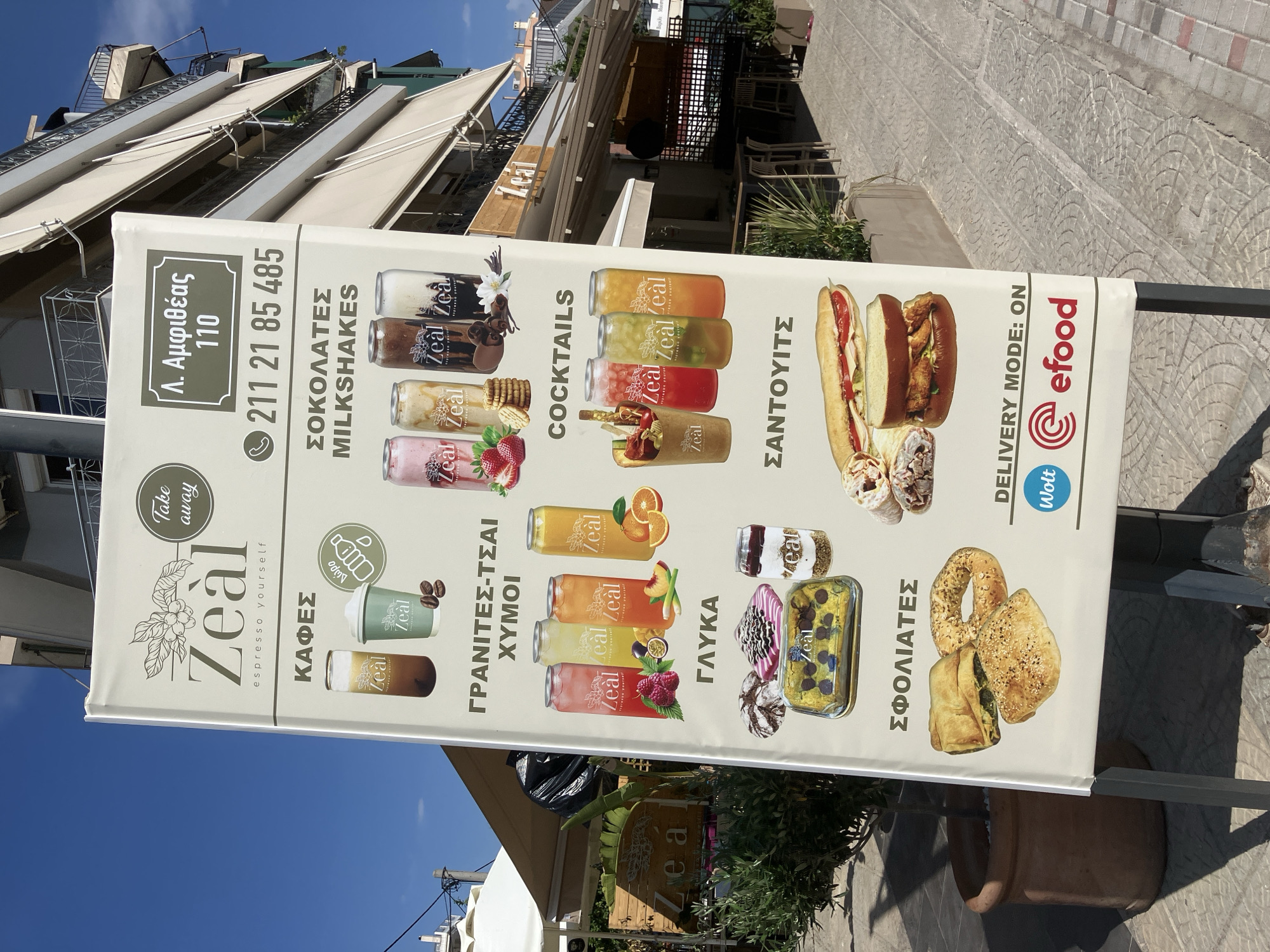 This is a photograph of a street-level advertisement sign for a cafe named "Zeal" which offers various drinks and food items. The sign is predominantly white with colorful images and Greek text.
The subjects on the sign include images of milkshakes, cocktails, juices, coffee cups, sandwiches, pastries, and sweet treats. The text indicates categories such as "MILKSHAKES," "COCKTAILS," "JUICES," "COFFEE," "SWEETS," and "PASTRIES." There is also contact information, including a phone number (211 21 85 485) and an address (Λ. Αμφιθέας 110). The sign also promotes delivery services with logos for "Wolt" and "efood."
The scene is set outdoors, likely on a sidewalk or in front of a business. The background shows a building with balconies and awnings, suggesting an urban or commercial setting. The sky is blue and clear, indicating a bright, sunny day. The sign is mounted on a metal stand and appears to be oriented vertically. The image is taken from a slightly elevated angle, looking up at the sign and the building behind it. The overall impression is one of a vibrant establishment ready to serve customers.