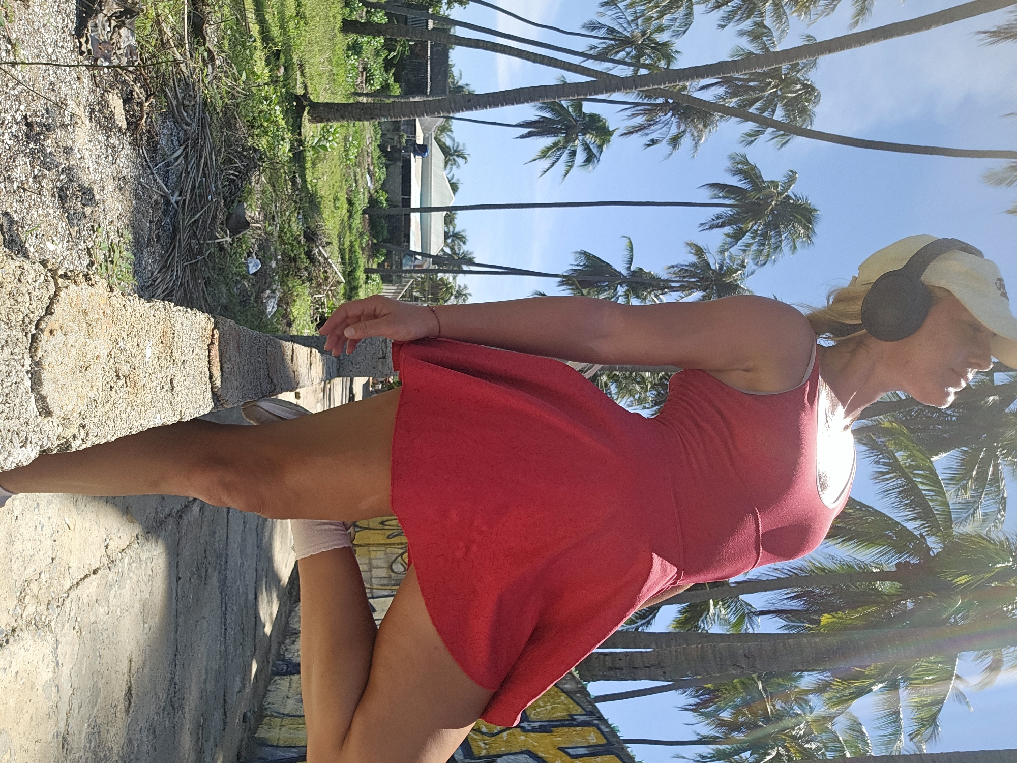 The image depicts a woman standing with one leg extended, leaning against a low concrete wall. She is wearing a red dress, a white baseball cap, and headphones. The background features numerous palm trees, a building with a light-colored roof, and a wall covered in graffiti. The scene appears to be an outdoor environment, possibly in a tropical or subtropical location. The woman's pose and the vibrant colors suggest a casual or recreational setting. The specific location of Phu Thuy Ward, Vietnam, is not directly evident from the visual information alone, but the presence of palm trees and the architectural style of the building are consistent with a Southeast Asian environment.