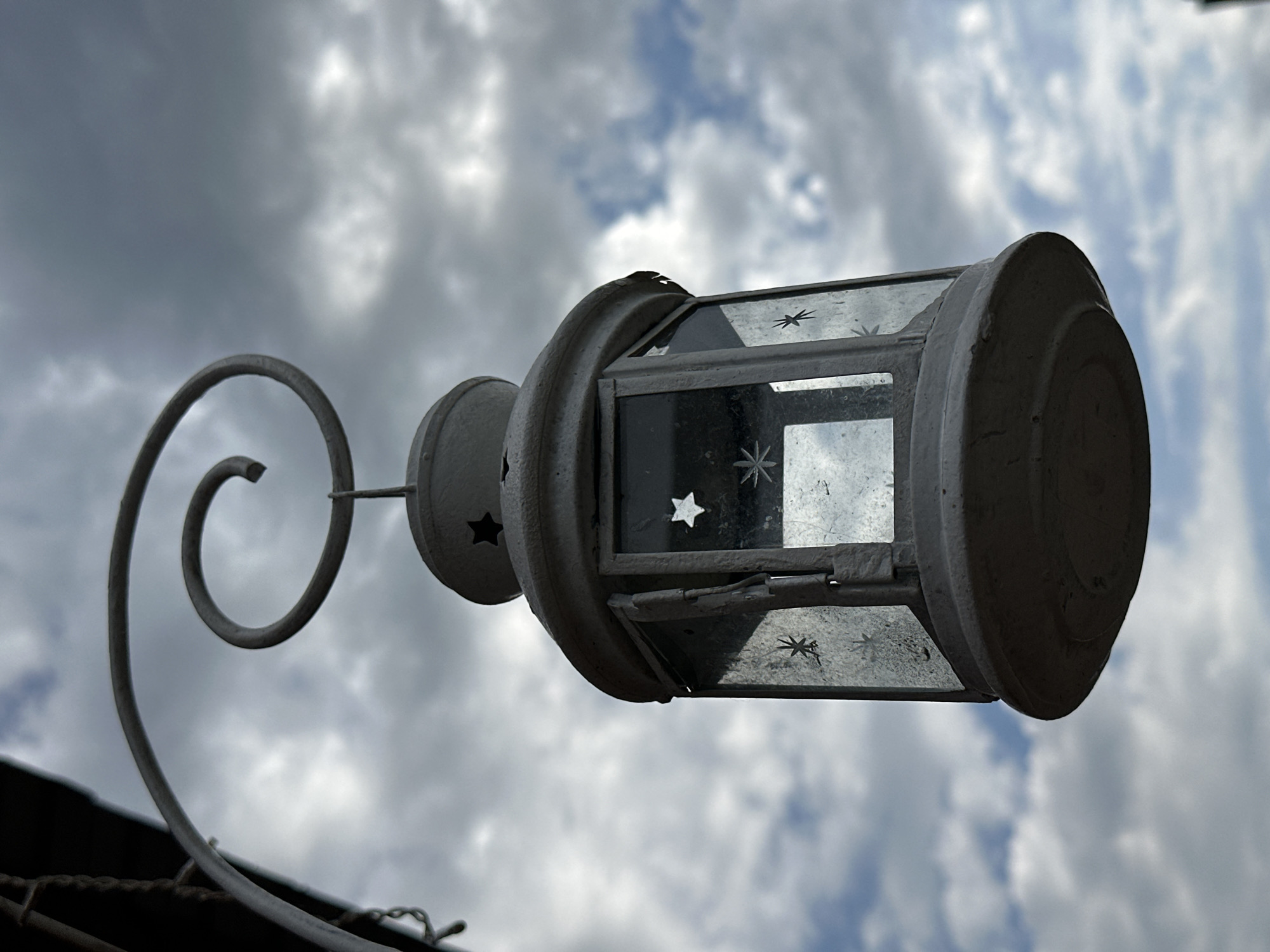 A low-angle, outdoor, eye-level shot captures a weathered, white, metal lantern hanging from a curved hook against a cloudy sky. The lantern is octagonal with glass panels that are smudged and slightly opaque, featuring etched star designs. The top of the lantern has small cut-out star shapes. A decorative swirl is part of the hanging mechanism. The background consists of a dramatic sky with a mix of dark gray and light blue clouds, suggesting overcast or stormy weather. The scene appears to be in an outdoor setting, possibly a garden or porch, but no specific urban landmarks are visible.