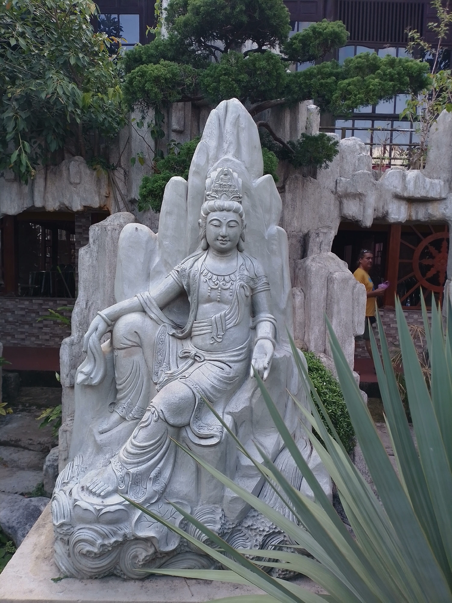 A serene outdoor setting in Thôn 5, Vietnam, features a white stone statue of a seated deity, adorned with intricate carvings and seated on a lotus-like base. The statue rests against a backdrop of fabricated stone cliffs, embellished with greenery. Architectural structures, with large windows and what appears to be a wheel-shaped window decoration, frame the scene. In the background, a person in a yellow shirt is present. Tall, blade-like plants partially obscure the foreground.