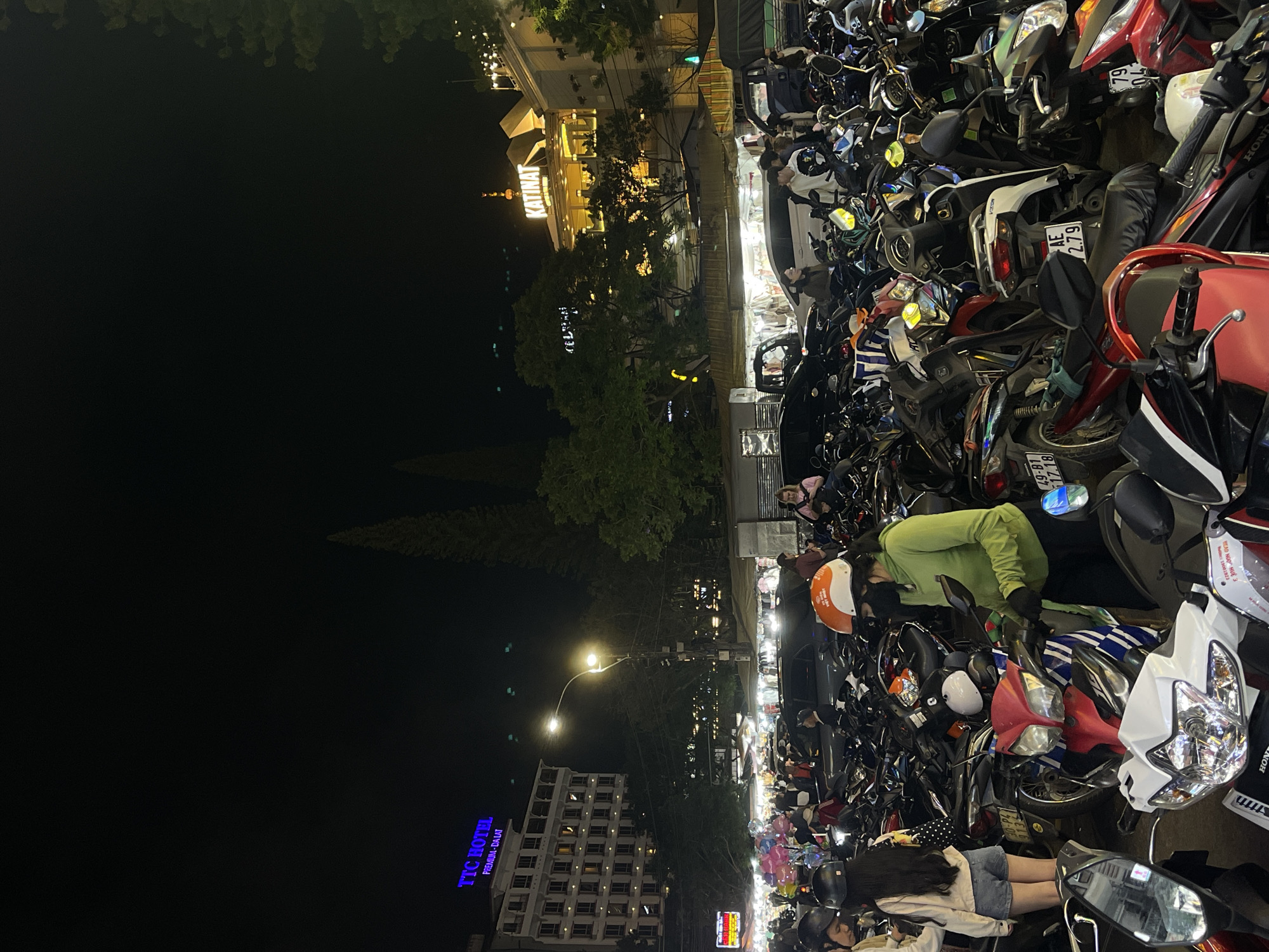 This nighttime view of Da Lat, Vietnam, shows a densely packed area with many parked motorbikes. People are present amongst the vehicles. A sign for the "TTC Hotel Premium - Da Lat" is visible. The "KATINAT" building is illuminated in the background. Streetlights illuminate the scene. Some of the motorbikes display license plates.