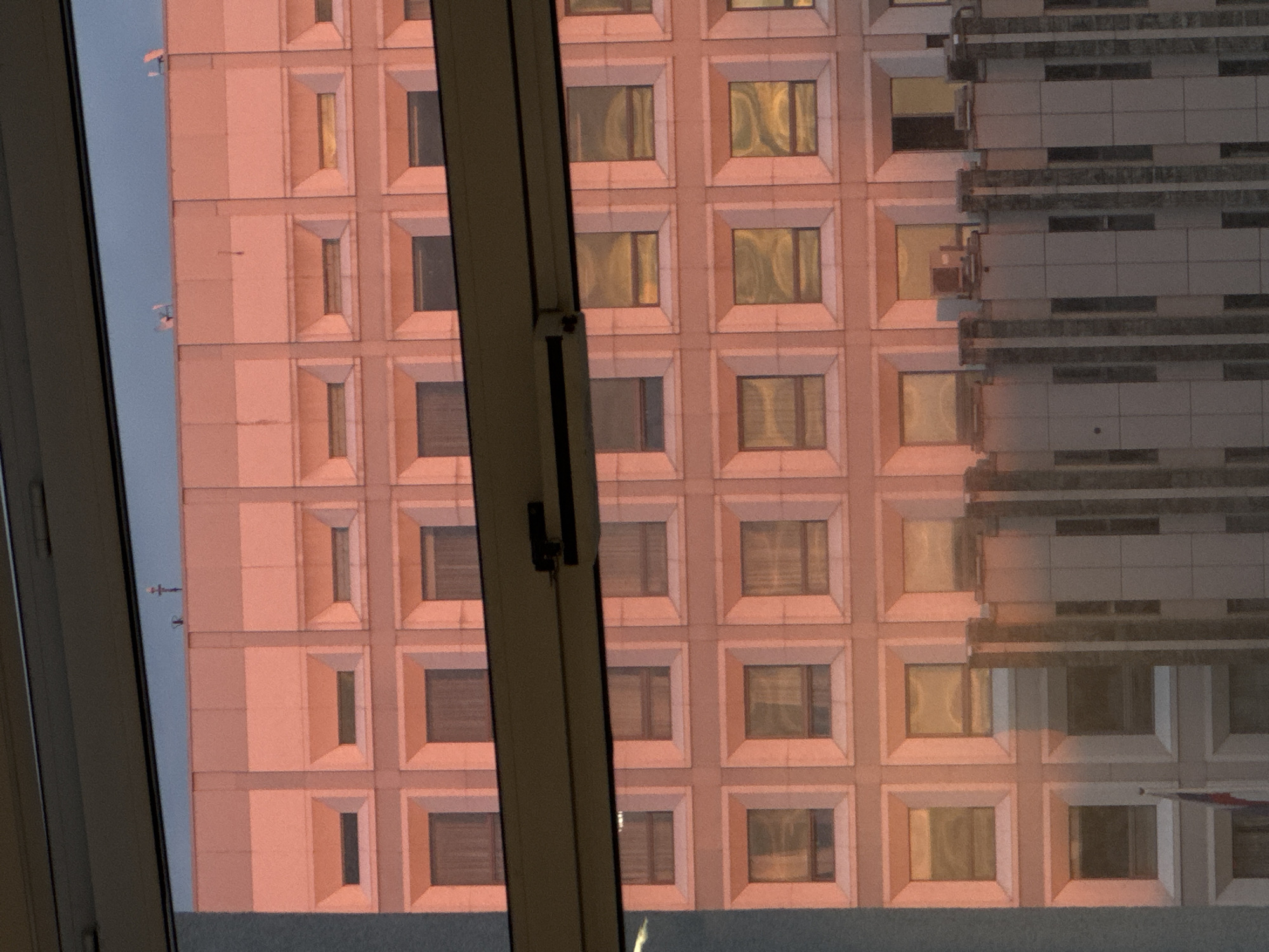 The image shows the exterior of a building with a regular grid of windows, viewed through a window frame. The building has a pink hue, possibly due to the lighting conditions. Part of the building is partially obscured by a section that appears to have a layered design. The view is partially framed by the window and its mullion.