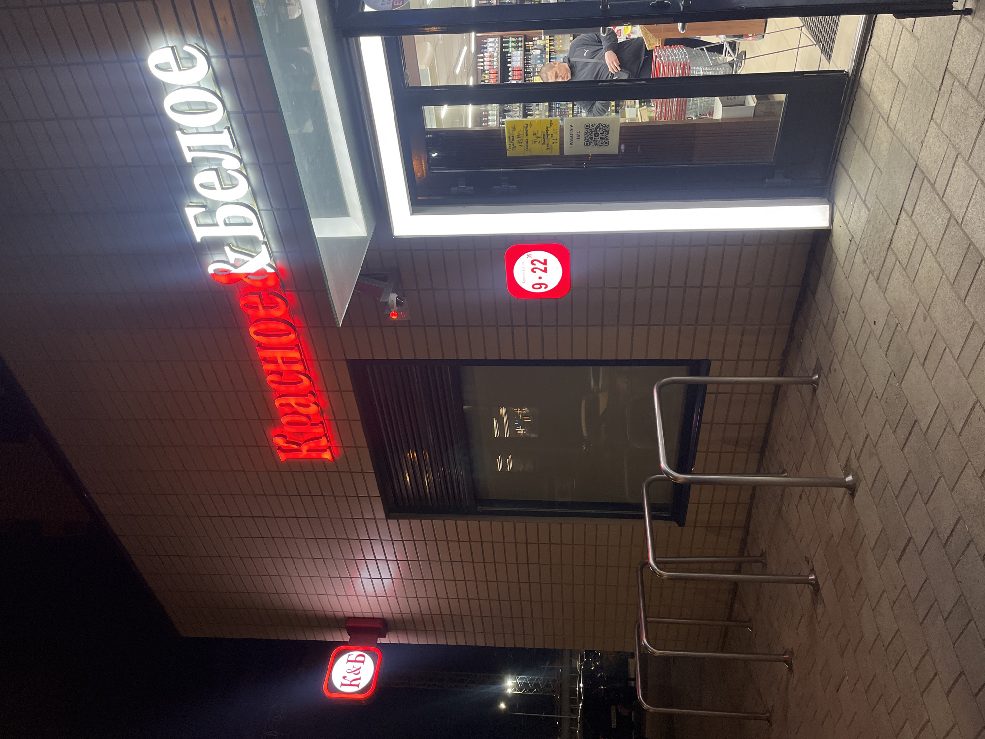The image depicts the brightly lit exterior of a store at night in Kudrovo, Russia. The building features a textured, light-colored brick facade. The dominant feature is a large, illuminated sign above the entrance, displaying the store's name: "Красное & Белое" (Krasnoye & Beloye, meaning "Red & White"), with "Красное" in red glowing letters and "& Белое" in white.
To the lower left of the main sign, a smaller red square sign with the abbreviation "К&Б" (K&B) is visible. Near the glass entrance door, another red square sign displays the operating hours, "9-22" (9 AM to 10 PM). A security camera is mounted on the wall above this sign.
Through the glass entrance door, shelves stocked with bottles, likely alcohol, are visible, along with shopping carts. One person, wearing a dark jacket, can be seen inside the store. A yellow notice inside the door displays text such as "Оплата по..." (Payment by...), "Картой" (Card), and "Наличными" (Cash), alongside a QR code. Another sign inside reads "РАБОТАЕМ ДЛЯ ВАС" (We work for you).
To the right of the entrance, four stainless steel bicycle racks are installed on the paved ground, which consists of light-colored bricks. The scene is illuminated by the store's artificial lighting and signs, contrasting with the dark night sky. The overall impression is that of a retail establishment open for business.