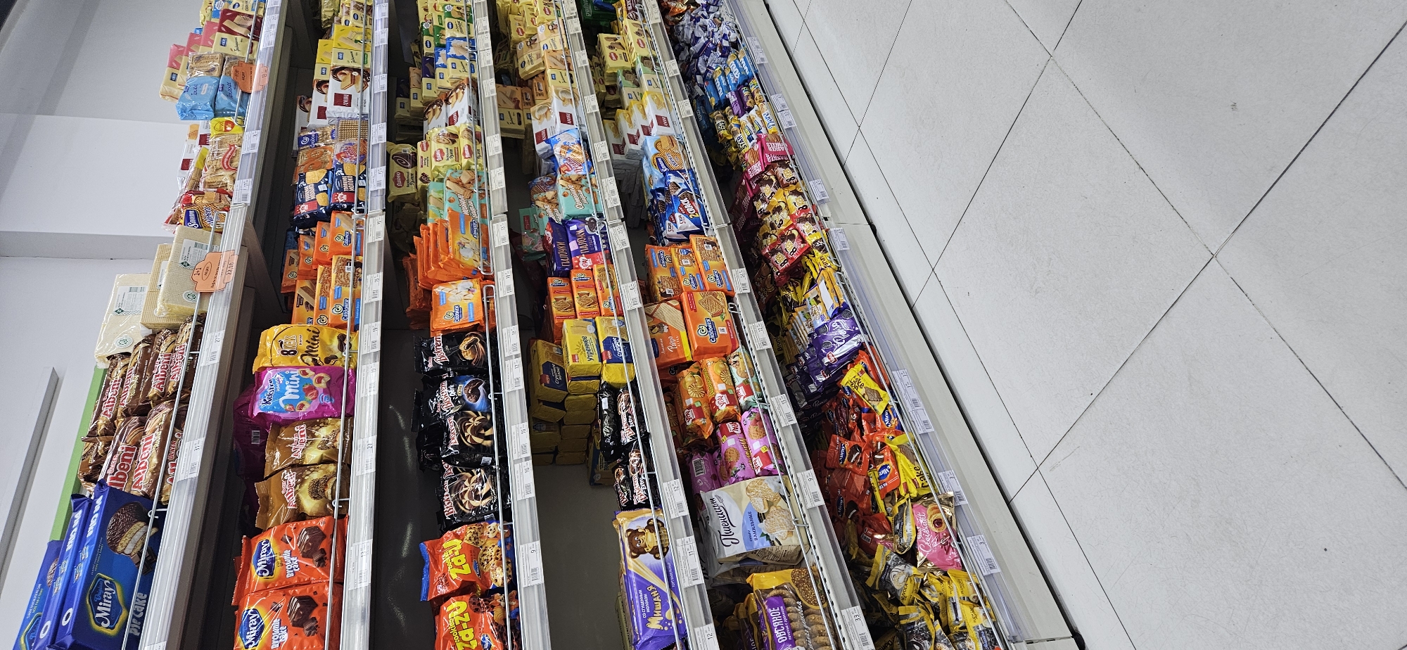 This is a medium-angle, eye-level, full-shot interior photograph of a supermarket aisle, specifically showing shelves stocked with various packaged confectionery items. The shelves are metallic and white, and they are illuminated by overhead fluorescent lighting, casting some reflections on the products. The products are predominantly boxes and bags of cookies, wafers, and other sweet snacks. They are neatly arranged in rows and stacks on multiple shelves, with some prices displayed on small white labels. The overall impression is one of abundance and variety. The floor is tiled with light-colored square tiles, some showing minor scuff marks. No people are visible in the frame. The background is out of focus, but appears to be the continuation of the supermarket interior. It is not possible to determine the specific location within Dushanbe, Tajikistan from the visual information provided.