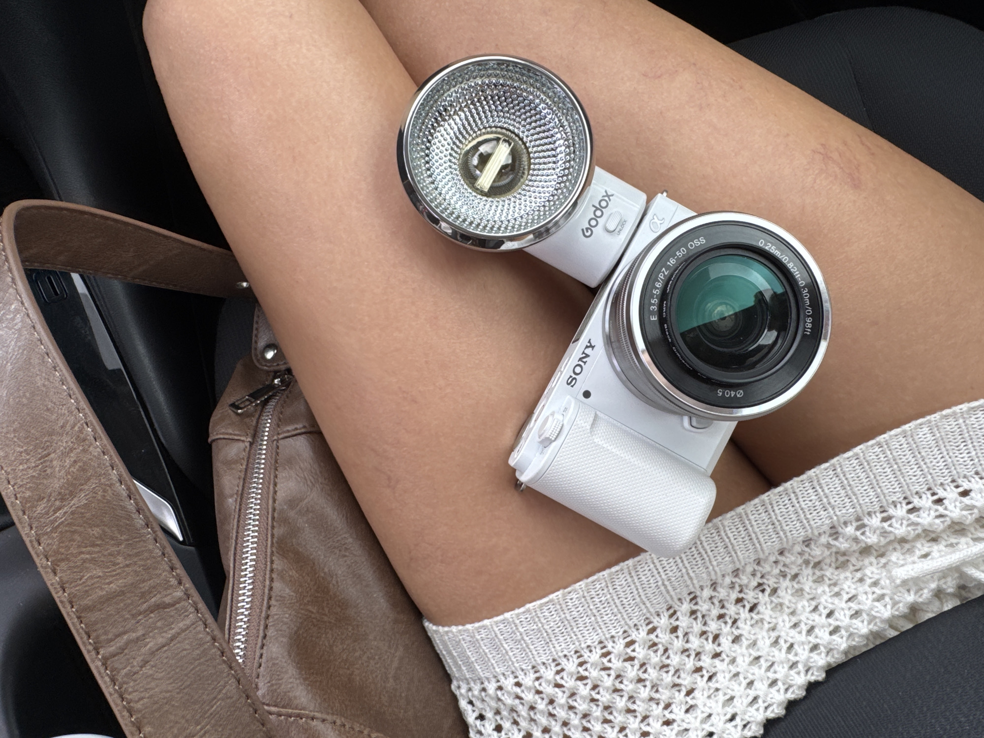 Here is a detailed description of the image:
**Scene Description:**
The image presents a close-up view of a woman's lap in what appears to be the interior of a car. A white Sony camera with a Godox flash attachment rests on her bare thigh, covered partially by a white knitted garment. A brown leather handbag with a visible strap and zipper is positioned to the left. The background shows the interior of a vehicle, including a black car door and a part of the car seat. The camera is the focal point of the image, with details visible in the lens and the flash attachment.
**Objects:**
* **Sony camera:** White, with a visible lens marked "E 3.5-5.6/PZ 16-50 OSS".
* **Godox flash attachment:** Attached to the camera, silver reflector visible.
* **Brown leather handbag:** With a visible strap and silver zipper.
* **White knitted garment:** Covering part of the thigh.
**People:**
* The primary subject is a portion of a woman's body (thigh and lap).
**Location:**
* Interior of a car. The location in the city of Palm Springs, United States of America cannot be confirmed from the image.