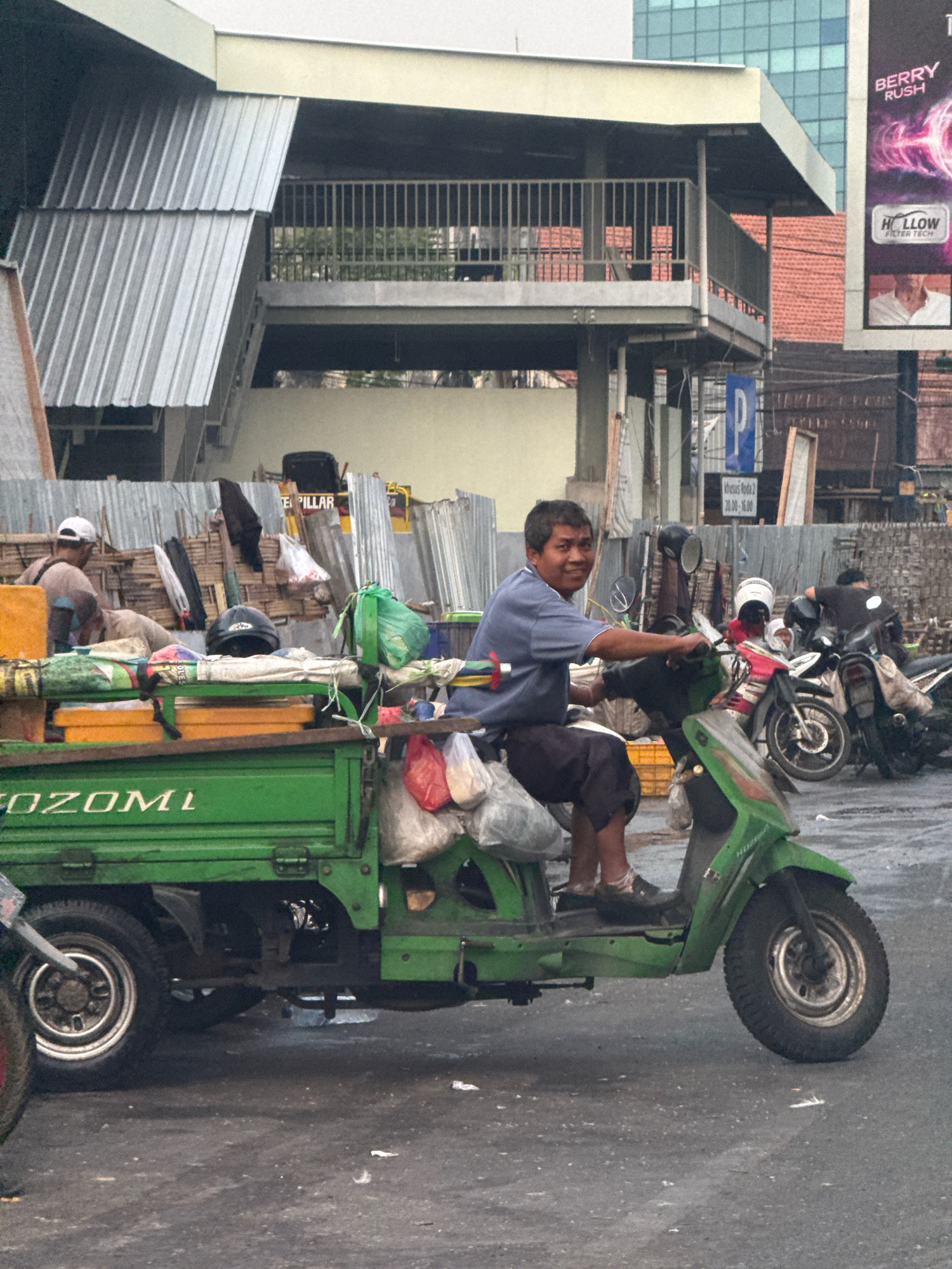 A man, appearing to be of Asian descent and in his late 30s to 40s, is seated on a green three-wheeled vehicle, resembling a motorcycle with a cargo bed, in what appears to be a street scene in Surabaya, Indonesia. He is wearing a blue short-sleeved shirt and dark shorts. The vehicle is loaded with various items, including what seem to be plastic bags, containers, and possibly food items, some of which are covered by green tarpaulin. A black helmet rests on the cargo bed behind him.
In the background, other motorcycles are visible, some parked and others with riders. Behind the vehicles, there is a structure with a corrugated metal roof and fencing. A billboard with text reading "BERRY RUSH" and "HOLLOW FILTER TECH" is also visible. Further in the background, a modern building with a glass facade can be seen, along with other buildings with red tile roofs. The ground is paved, and the lighting suggests it is daytime. The overall scene depicts a busy urban environment.