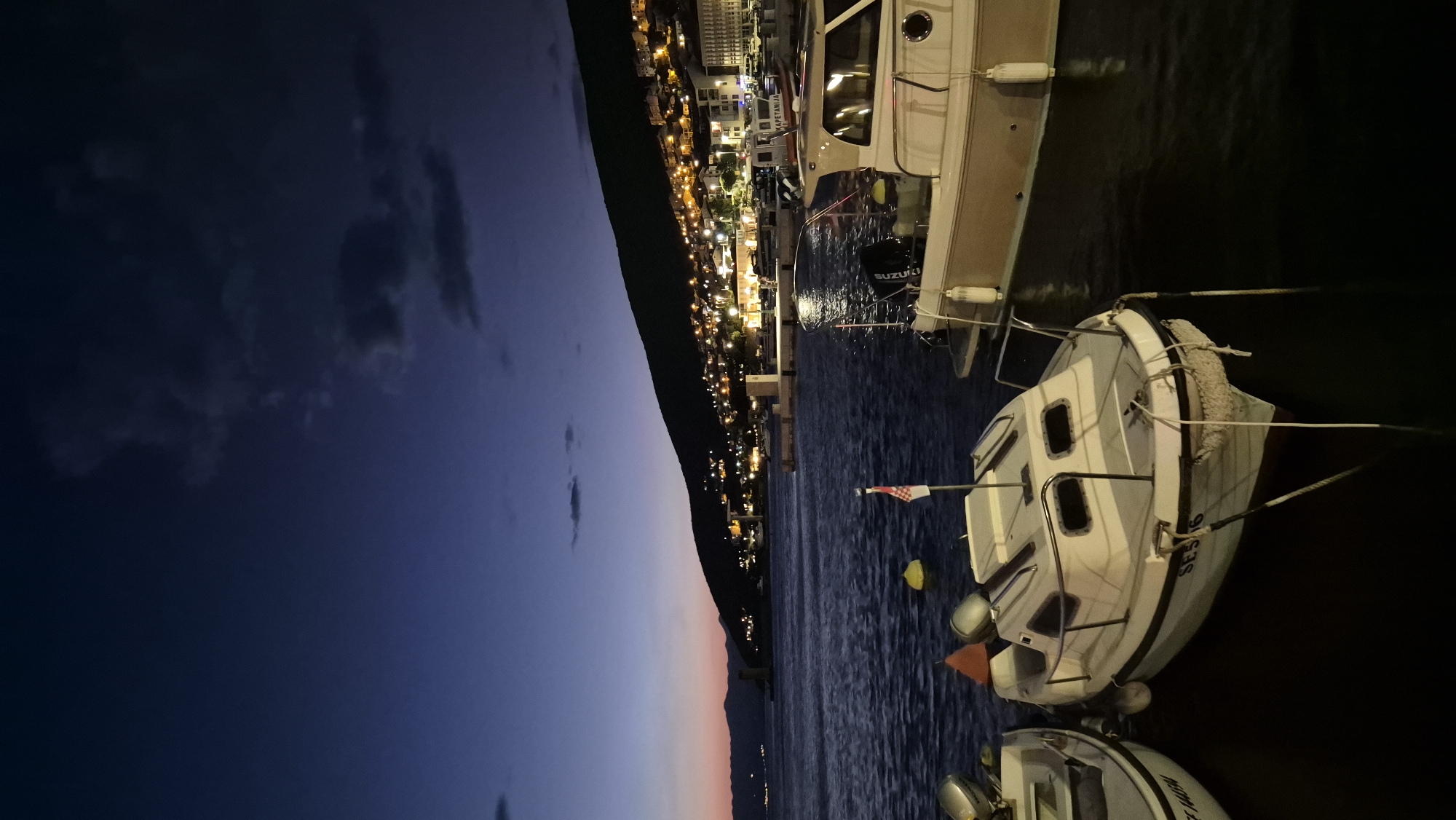 The image depicts a harbor scene at dusk, with a town illuminated on a hillside overlooking the water. In the foreground, two small boats are moored, one white with black trim and a red and white flag. A larger boat, light beige with a dark roof, is docked to the right. The water is dark and slightly rippled. The sky transitions from a deep blue at the top to hues of orange and pink near the horizon. The town's lights twinkle on the slope of the hill, creating a warm glow against the darkening sky. The overall impression is one of a peaceful evening in a coastal settlement.
