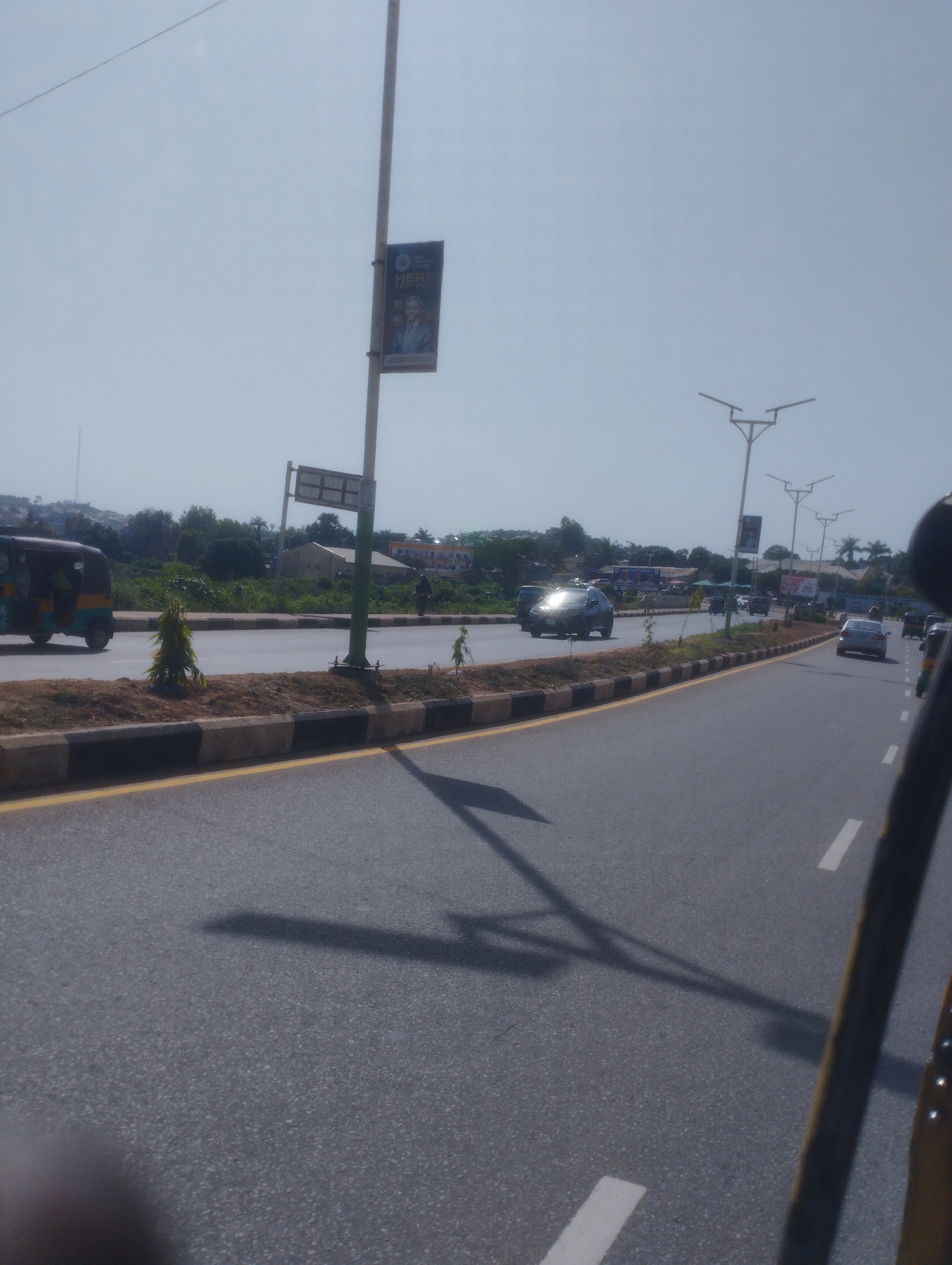 The image depicts a street scene in Jos, Nigeria. A multi-lane asphalt road is visible, with yellow lines marking the lanes. Along the side of the road, there is a curb with black and white stripes, followed by a dirt median where small trees and plants are growing. The scene is bathed in bright sunlight, casting shadows on the road.
In the background, there are buildings and dense vegetation. Several vehicles are present on the road, including a blue and yellow auto-rickshaw on the left, a dark-colored sedan in the middle, and other cars further down the road. There are also streetlights visible, some with modern designs. A tall pole with a banner is also present, featuring a portrait of a man and text that is partially visible. A few individuals can be seen on the roadside. The sky is clear and pale blue.