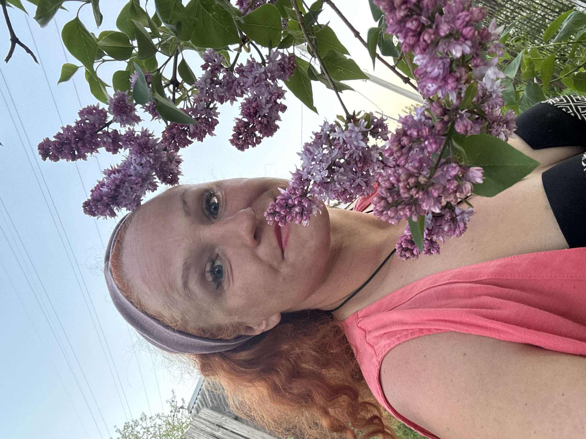 A woman with red hair and a headband is shown from a low angle looking up at the camera. She is wearing a pink shirt and a black top beneath it. She has fair skin and is wearing lipstick. Purple lilac flowers and green leaves are visible in the foreground, partially obscuring her face and upper body. The sky is visible in the upper left corner, suggesting an outdoor setting. A wooden fence is partially visible in the lower left corner, and power lines stretch across the sky in the background. The location is possibly Chaikovsky, Russia.