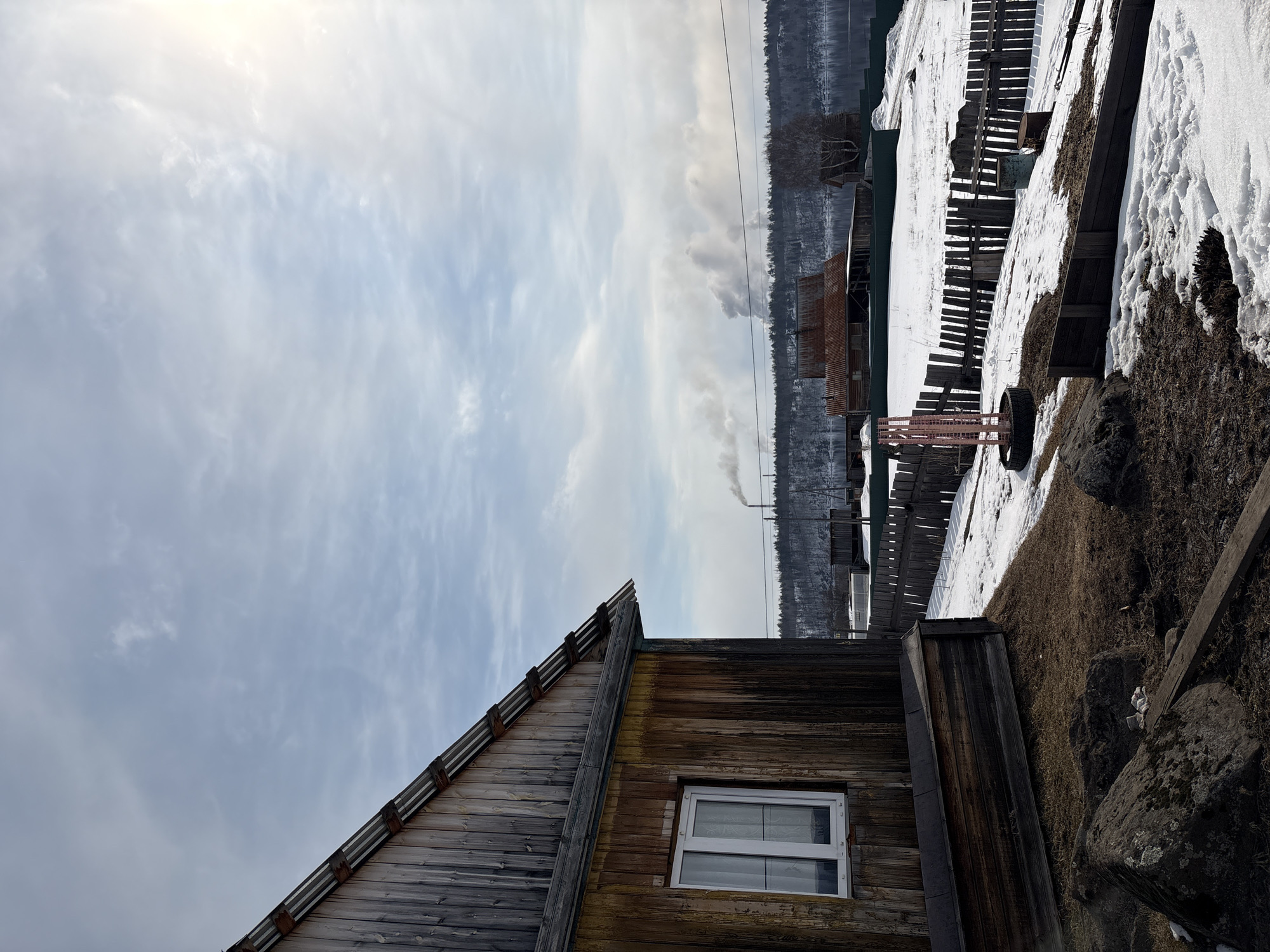 This landscape photograph captures a quiet, wintry or early spring scene in a rural or semi-rural area of Nevon, Russia.
In the foreground, the side and roofline of a weathered wooden house dominate the lower left. The house's facade features horizontal wooden planks in varying shades of gray, brown, and some yellowed areas, suggesting age and exposure. A single, white-framed, multi-pane window is visible on its side. The steeply pitched wooden roof also shows signs of wear. To the right of the house, the ground transitions from patches of dry, brown grass and dark soil to areas covered in snow and large rocks.
The midground features significant snow cover interspersed with bare ground. A rustic wooden fence, partially buried in snow, extends diagonally across the scene. Various weathered wooden structures, possibly sheds or parts of fences, and a large tire are scattered on the snow-dusted terrain. Further back, another older wooden building with a rusty corrugated metal roof is partially visible.
In the background, a dense line of dark evergreen trees forms a forest, stretching horizontally across the image, likely indicating a distant hillside or riverbank. Above the tree line, a faint plume of white smoke rises into the overcast sky from an unseen source, possibly a chimney or a distant industrial facility, located towards the right.
The sky, which fills the upper portion of the image, is mostly overcast with thin, wispy clouds scattered across a light blue-gray expanse. A bright spot in the upper left corner suggests the sun's position, obscured by clouds. The time of day appears to be daytime, likely morning or early afternoon, given the diffuse but bright light. The weather is cold, evidenced by the snow and overcast sky. No human activity or visible text is present, contributing to the serene, slightly stark atmosphere.