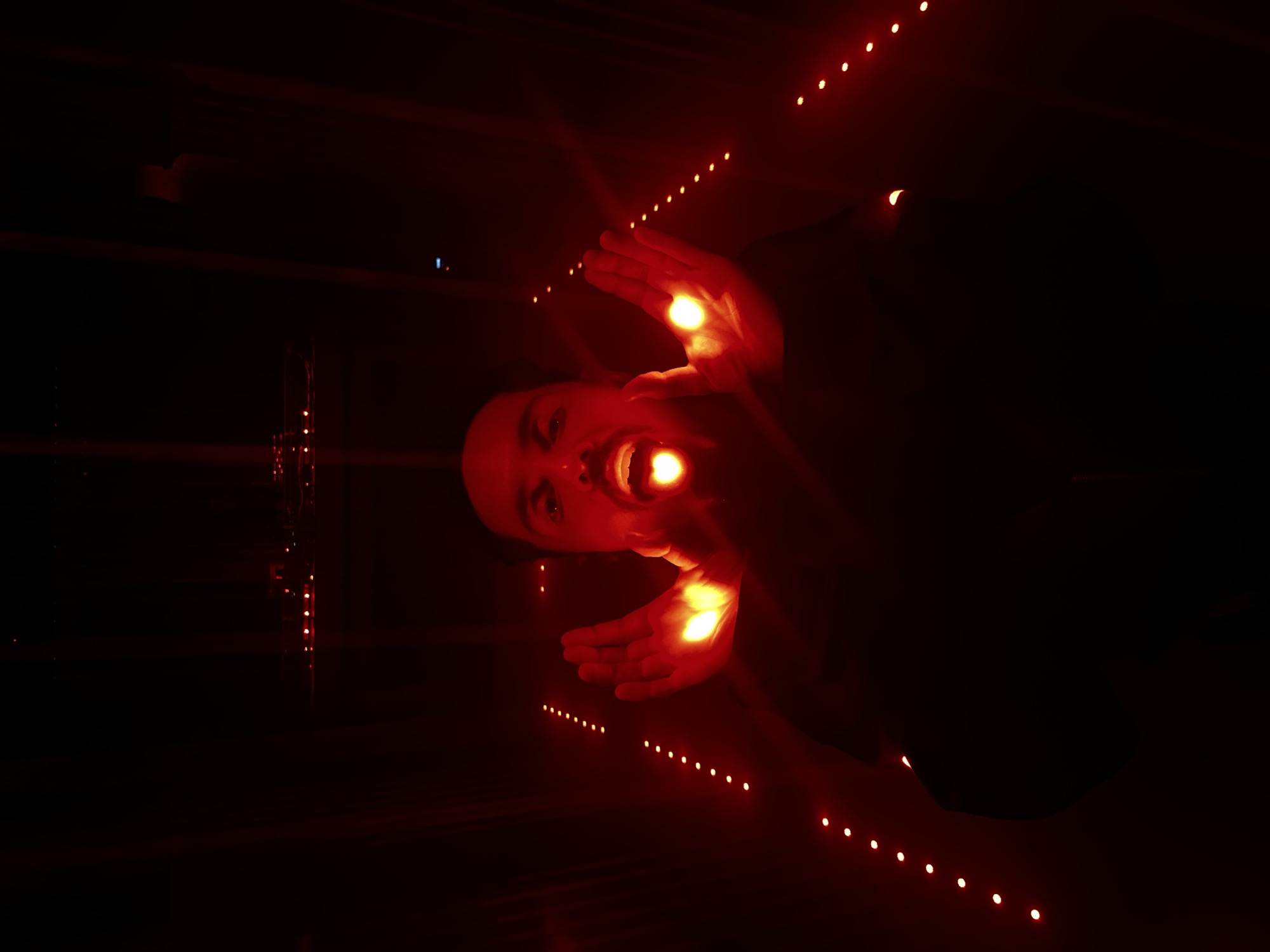 The image, presented with a vertical orientation where the subject's head is on the left, depicts a person's head and hands dramatically lit against a dark background. The central figure, seemingly looking upwards or lying down, has a yellow-orange glow emanating from their slightly open mouth and from the palms of both hands, which are held near their face. Their eyes are open and directed towards the viewer, conveying an intense or focused expression.
The background is profoundly dark, providing a stark contrast to the bright lights. Multiple parallel rows of small, evenly spaced, glowing red lights recede into the distance, creating a strong sense of perspective and depth. These linear arrays of red lights suggest an enclosed space, such as a long corridor, tunnel, or an art installation with immersive lighting.
The activity appears to be an artistic pose or performance, with the person seemingly "breathing" or "holding" light. The overall atmosphere is mysterious, theatrical, and dreamlike, emphasizing the interaction between the person and the glowing elements. The scene is set in a dark environment, indicative of night or an artificially darkened indoor space. No specific location cues or visible text are present within the image.
