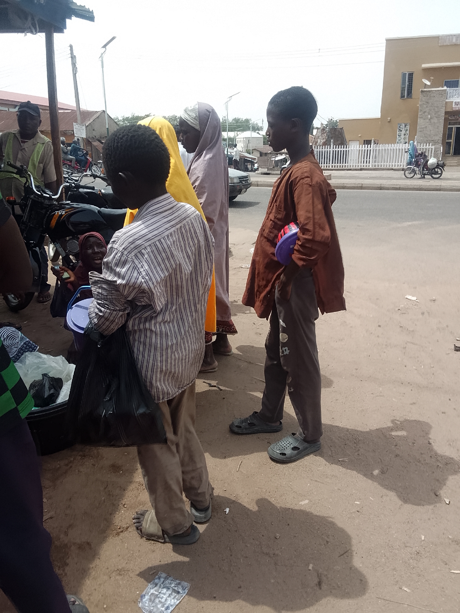 A daytime scene shows a group of people gathered in what appears to be a street in Pantame, Nigeria. A boy wearing a brown shirt and grey pants stands on the right, holding a purple and red object. Next to him are two people, one in yellow clothing and another in a lilac-colored headscarf. To the left is a boy in a striped shirt holding a plastic container. In the background, there is a motorcycle and a man standing next to it. There is also a woman on the left, a vendor, and another woman in the background by the motorcycle. A building is visible across the road. The ground is bare dirt, and there are shadows.