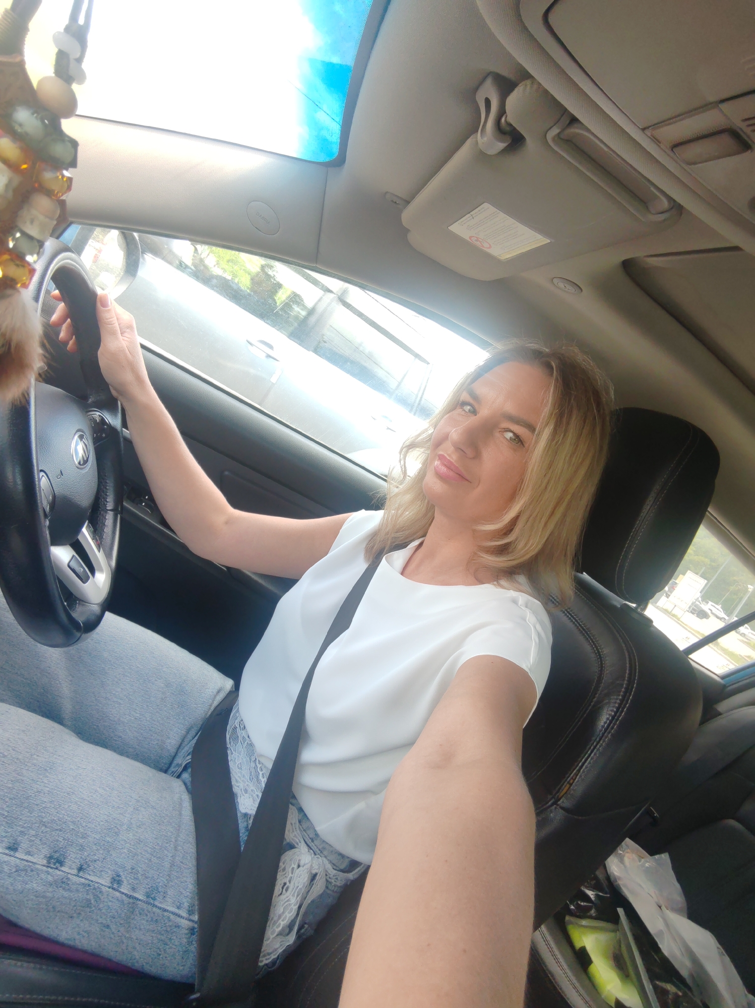 In Petropavlovsk-Kamchatsky, Russia, a woman with blonde hair and fair skin, wearing a white top and light blue jeans, is seated in the driver's seat of a car. She is holding the steering wheel and looking towards the camera with a slight smile. The interior of the car, including the dashboard and seats, is visible. Sunlight streams in through the sunroof and windows, illuminating the scene. Outside the car, glimpses of other vehicles and trees can be seen, suggesting an urban or suburban environment. A beaded ornament hangs from the rearview mirror.
