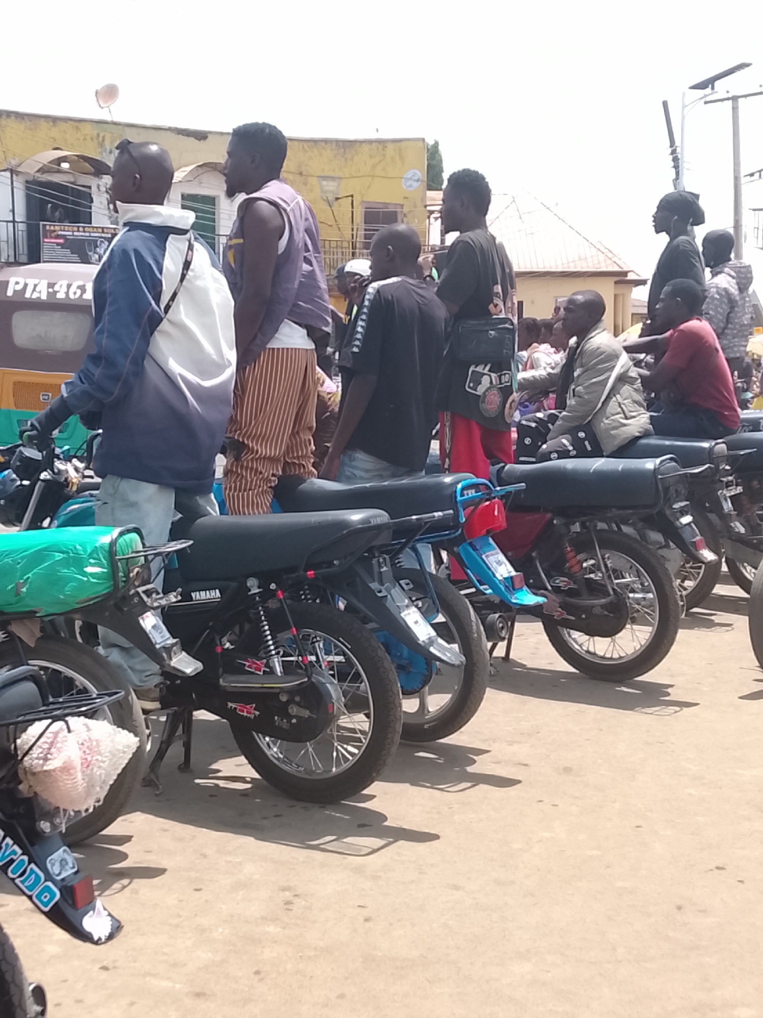 The image depicts a street scene in Gwong, Nigeria, with a line of parked motorcycles and a group of men standing nearby. The motorcycles, some bearing the Yamaha brand, are varied in color and condition. Several men are standing or sitting on the motorcycles, while others are standing on the street next to the motorcycles. A building is visible in the background, marked with the text "SARTECH & OGAR SOLU", the roofline suggests other buildings. The environment appears to be a sunny day.