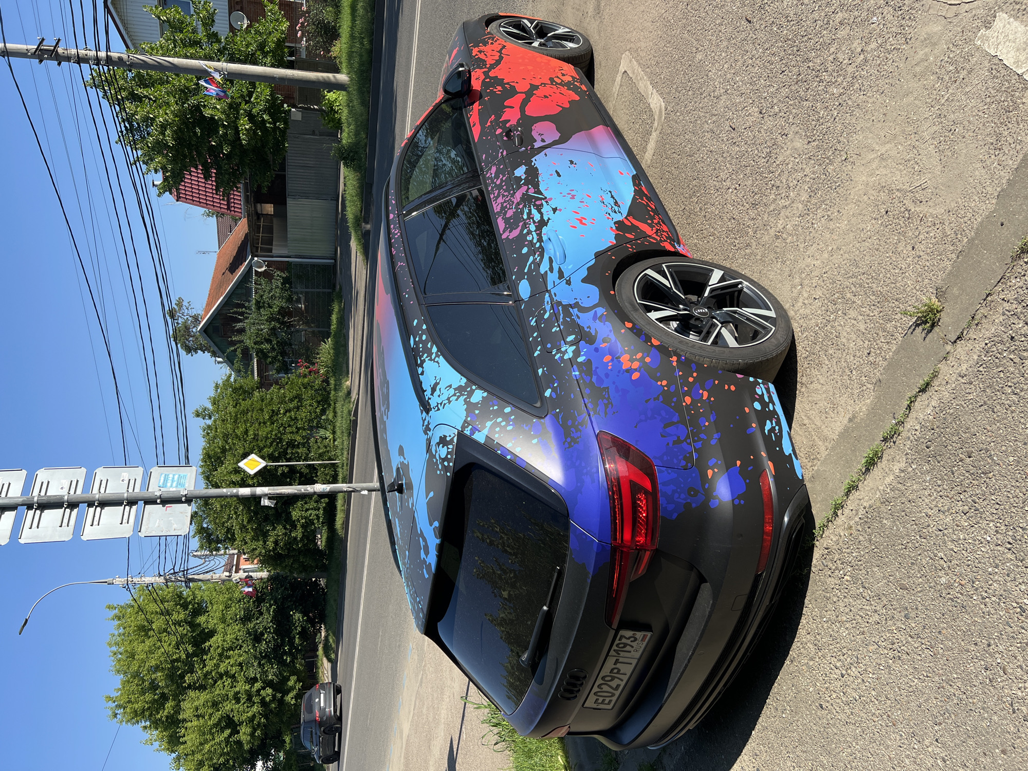 The image depicts an outdoor street scene under a clear blue sky, likely during daylight hours, in Krasnodar, Russia.
The central subject is a black Audi A4 Avant (wagon) parked on the side of an asphalt road. The vehicle features a custom vinyl wrap characterized by a black matte background adorned with irregular, paint-splatter patterns. The rear and roof sections of the car display predominantly blue and purple splatters, while the side transitions towards the front feature pink and red splatters. The Audi is equipped with multi-spoke black alloy wheels that have polished silver accents on the spokes. The rear license plate, "E029PT 193 RUS," is clearly visible, identifying the vehicle as registered in Krasnodar Krai, Russia. The car's windows are tinted.
To the left of the parked Audi, a two-lane asphalt road is visible with a white solid line marking the shoulder. A dark-colored SUV, possibly a Volkswagen Tiguan, is driving in the distance on this road. The Audi is parked on a paved area adjacent to the curb, which separates it from a strip of grass.
The environment is characterized by lush green vegetation. Large, mature deciduous trees with dense foliage line the left side of the road, and smaller trees and bushes are present on the right.
Various elements of urban infrastructure are present:
* A tall metal utility pole, equipped with a street light, stands on the left.
* Numerous black electrical or communication wires crisscross the upper portion of the frame, connected to utility poles.
* A signpost with four rectangular grey signs and a yellow diamond-shaped "priority road" sign is positioned near the center of the frame. The text on the grey signs is not legible.
* Another wooden utility pole is visible on the right, from which two small Russian national flags (horizontal white, blue, and red stripes) are affixed.
In the background, behind the trees and utility poles, several residential buildings with pitched roofs, some red-tiled and others with green or lighter colored roofing, are partially visible. The lighting suggests a sunny day, with shadows cast on the ground.
