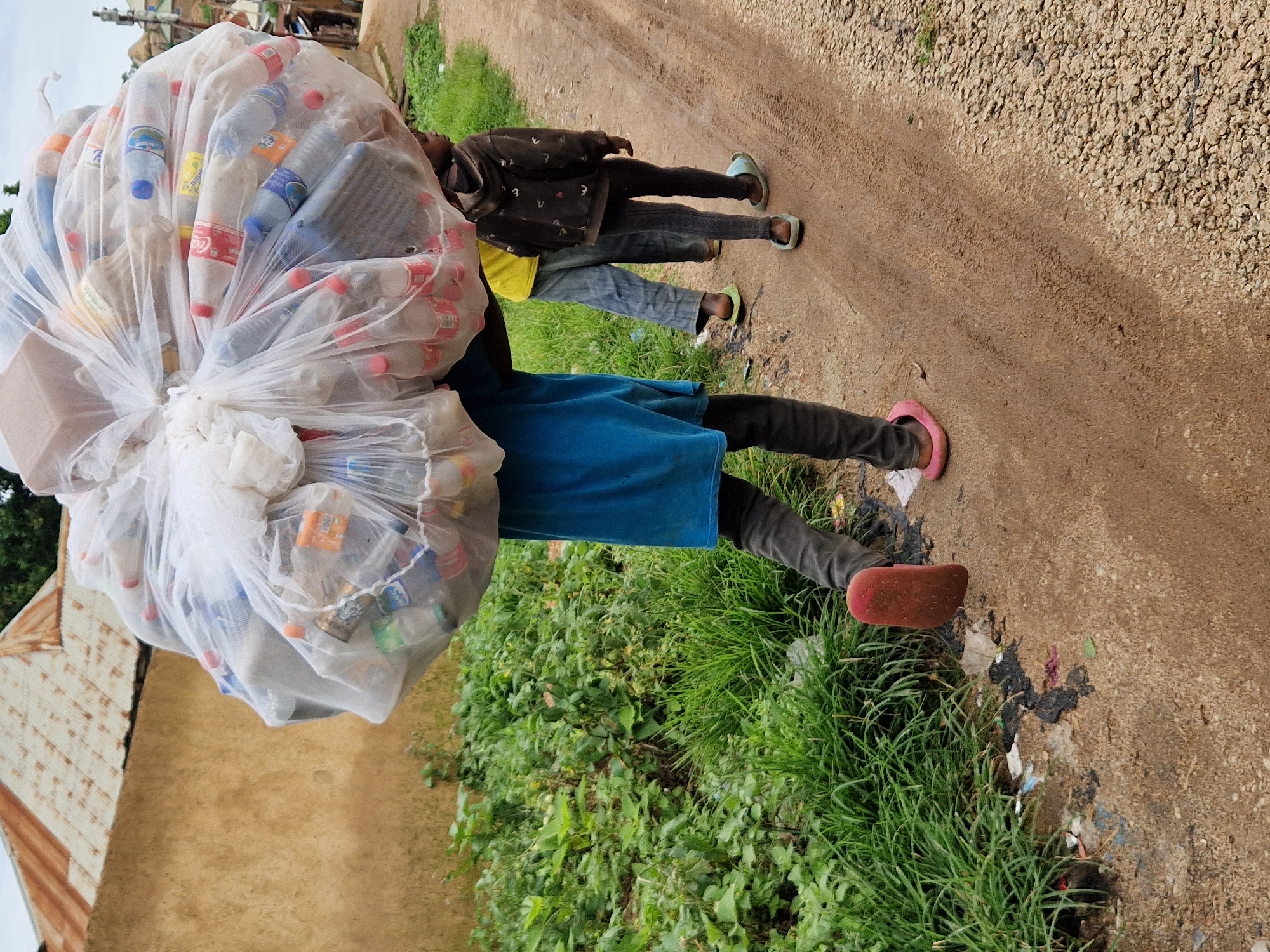 In the city of Unknown, Unknown, a child, whose body is obscured by a large, clear plastic bag overflowing with empty plastic bottles, carries the load on their shoulders. The child wears dark pants and a blue shirt. Behind them, another child, wearing a brown hooded sweatshirt with a patterned design and dark pants, walks on a dirt road. To the left of the road, dense green vegetation grows, with some scattered debris on the ground. In the background, a building with a rusty corrugated metal roof is visible. The road is a mix of dirt and gravel, with tire tracks imprinted on the surface. The sky is overcast.