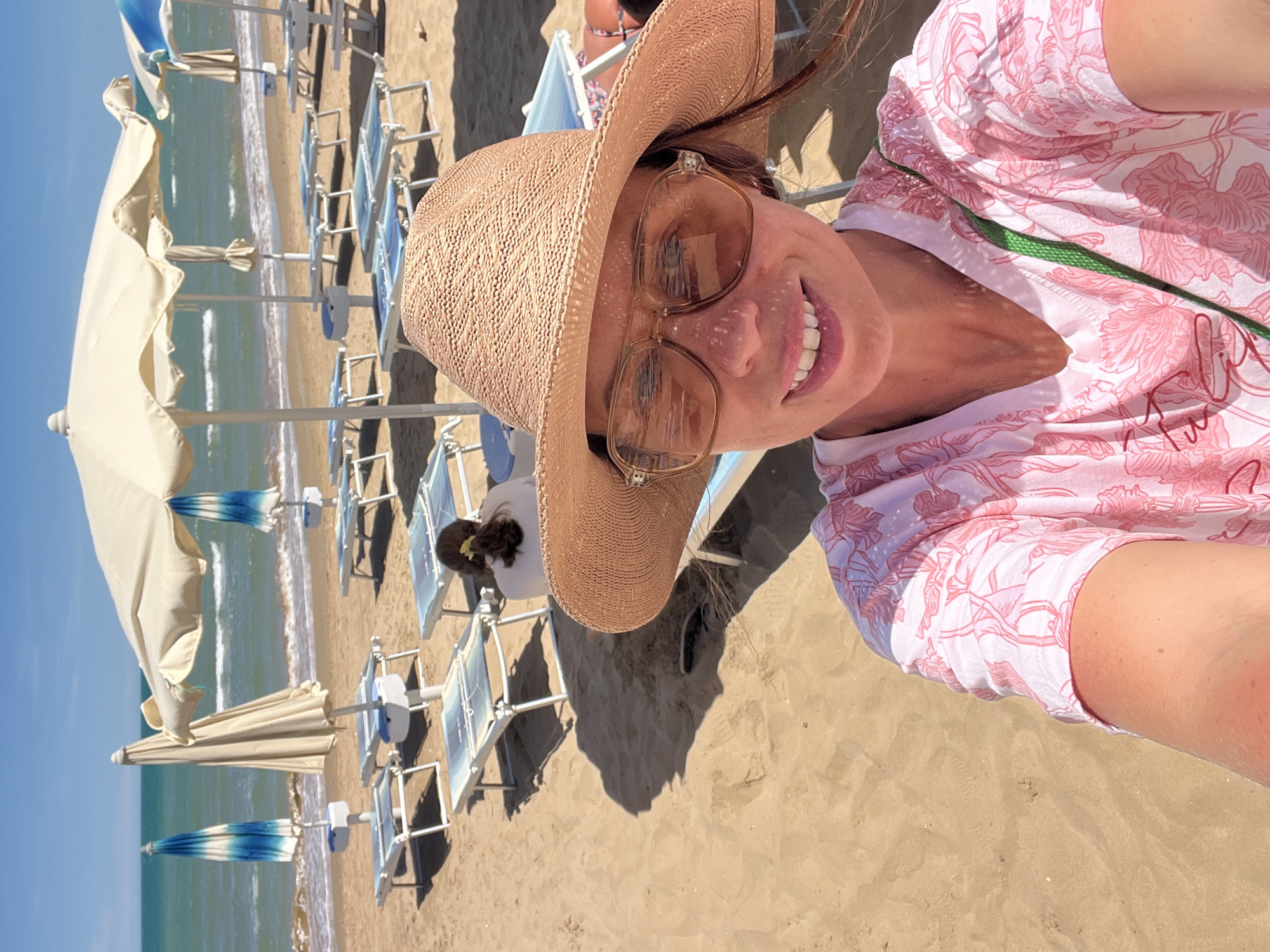 A woman takes a selfie on a sandy beach in Peschici, Italy, wearing a straw hat and oversized sunglasses. She has a pink floral-patterned shirt on and a green strap visible. Behind her, beach chairs and umbrellas are set up, with the ocean in the background. The sky is clear and blue.