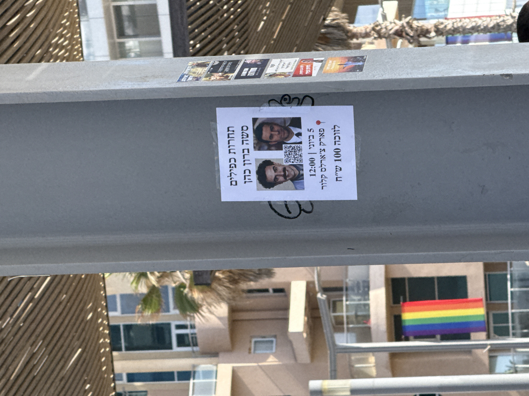 The image shows a grey metal beam with a flyer attached to it. The flyer advertises a Sasha Baron Cohen look-alike contest scheduled for June 5th at 12:00 in Charles Clore Park. The flyer features two images of people who resemble Sasha Baron Cohen and a QR code. Above the flyer are several other stickers. In the background, a building with a rainbow flag is visible, along with a palm tree and other buildings. This indicates the location is likely in Tel Aviv, Israel, near the beach.