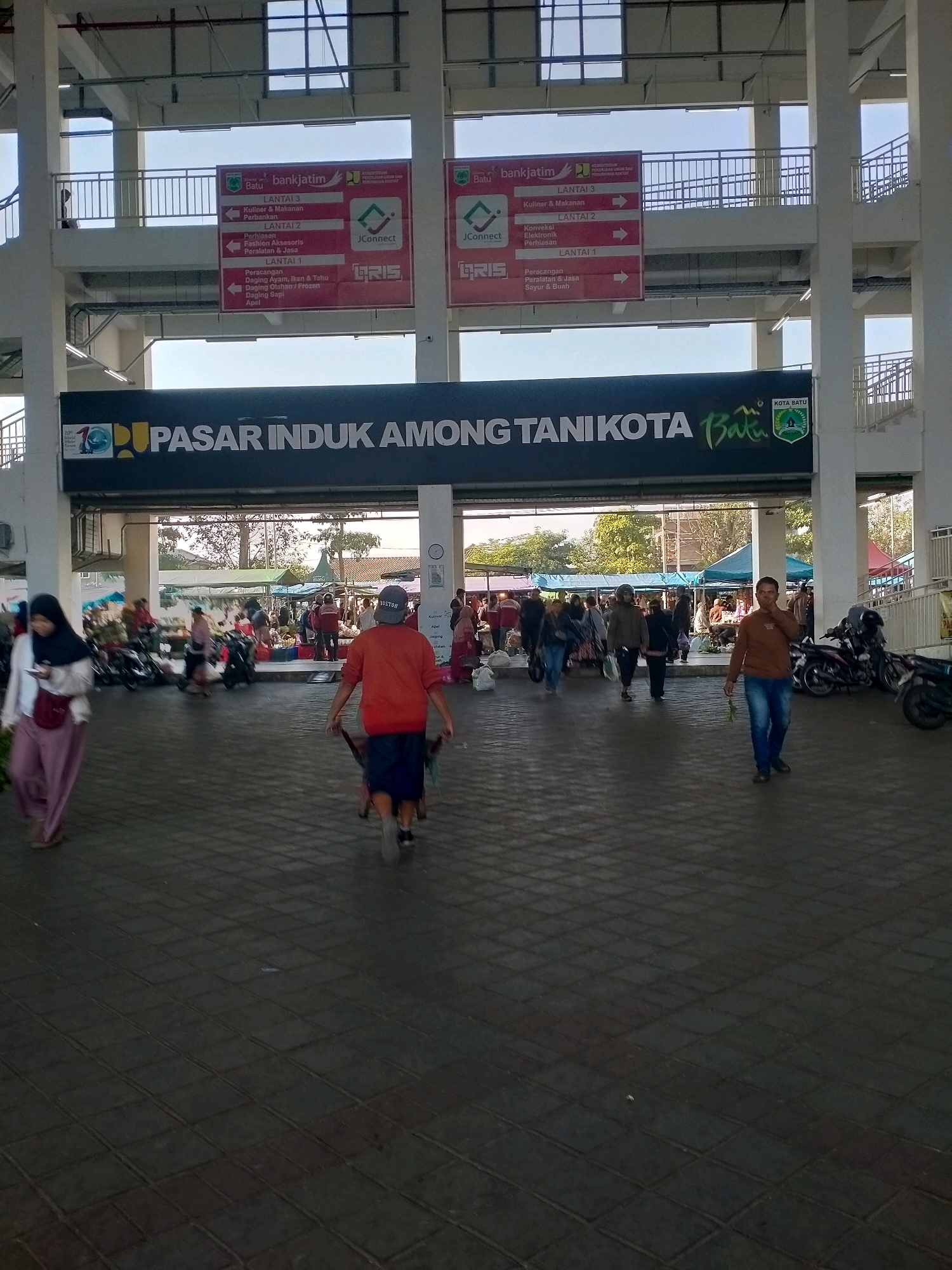 This is a photograph taken at the "Pasar Induk Among Tani Kota Batu" in Batu, Indonesia. The image captures a bustling market scene with people engaged in various activities. In the foreground, a person wearing an orange shirt and blue shorts is pushing a cart, walking away from the camera. To the left, a woman in a hijab and pink skirt is walking towards the camera, holding a mobile phone. Further into the market, several individuals are seen walking, some pushing carts, and others browsing. Tents and stalls are set up, indicating the presence of vendors selling goods. Motorcycles are parked in the background. Above the market entrance, a large black banner with white lettering reads "PASAR INDUK AMONG TANI KOTA Batu", with a city emblem on the right. Overhead, there are elevated walkways with railings and structural supports, typical of a large market complex. The lighting suggests it is daytime, with sunlight filtering through the open structure of the market.