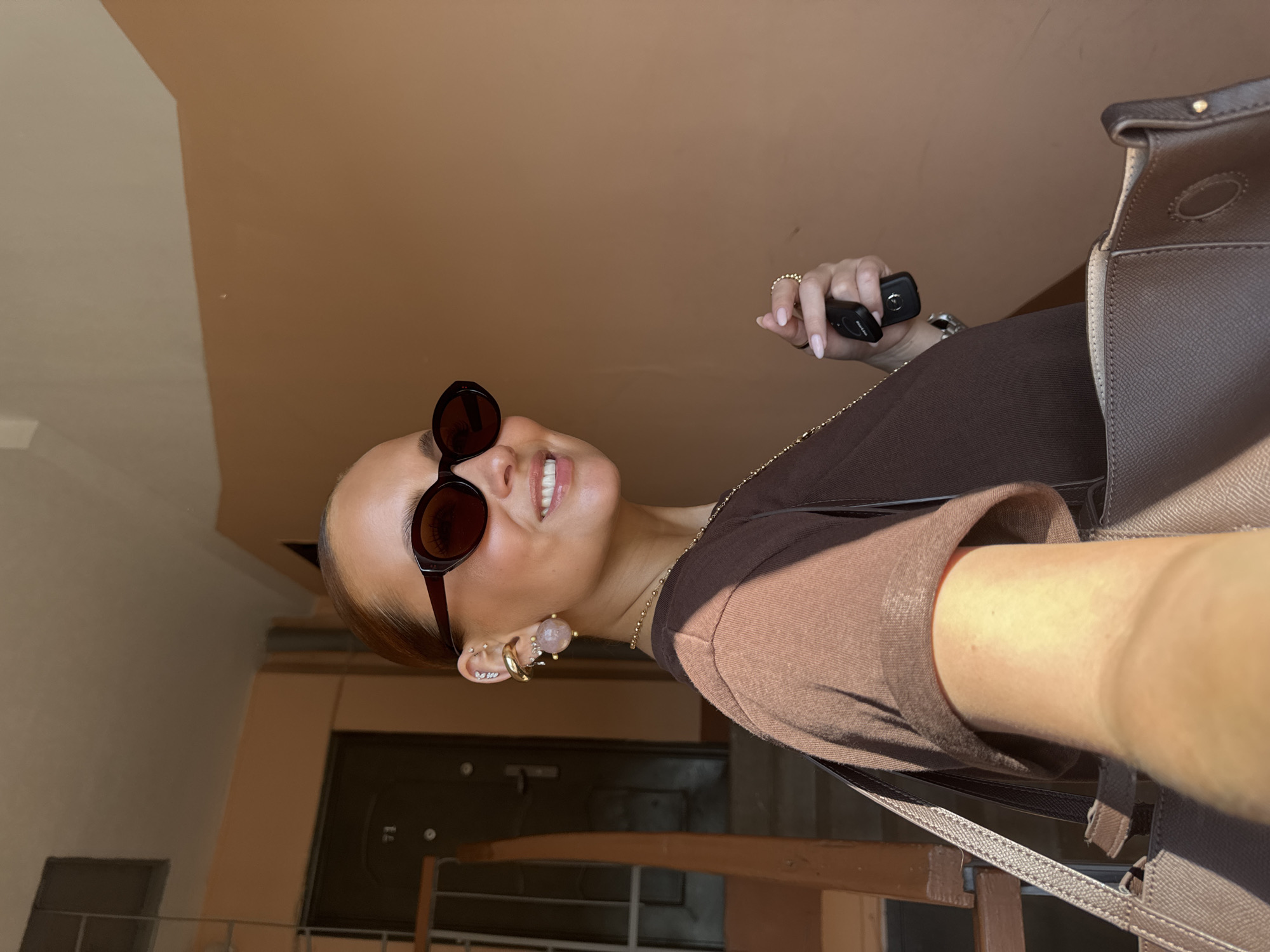 A woman is taking a selfie in a building. She is wearing sunglasses, earrings, and a brown shirt. She is holding a car key in her hand. The background shows a hallway with a door and a brown wall. Based on the architectural style and the information provided, the location is likely in a building in Chaikovsky, Russia.