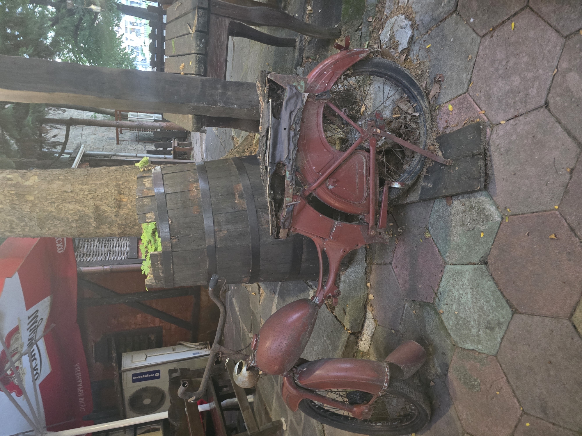 The image depicts an old, rusted red moped parked on a hexagonal paved surface in Plovdiv, Bulgaria. The moped is leaning against a tree trunk, and a large wooden barrel is situated next to it. In the background, a portion of a red umbrella is visible, with text that appears to be in Cyrillic script. A white air conditioning unit is mounted on a wall to the left of the moped. The overall scene suggests an outdoor seating area, possibly part of a cafe or restaurant, with a rustic and weathered aesthetic. Fallen leaves are scattered on the ground around the moped and barrel.