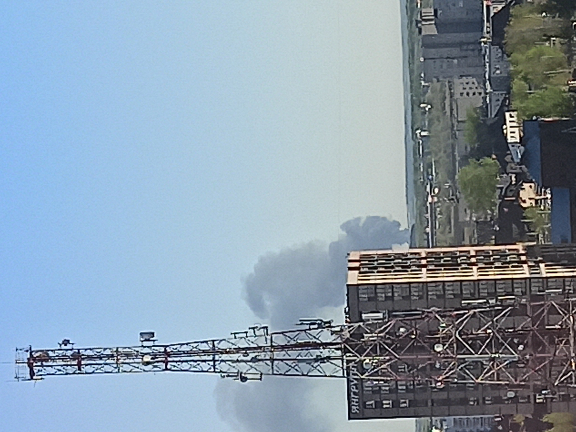 The image shows a large plume of dark smoke rising in the vicinity of buildings and an antenna structure in Кондратово, Russia. The smoke plume appears dense and widespread. The foreground features a metallic tower structure with various antennas and communication devices. A multi-story building is partially visible to the right of the tower. The backdrop includes a cityscape with buildings, greenery, and sky. The sky is clear with a slight blue gradient.