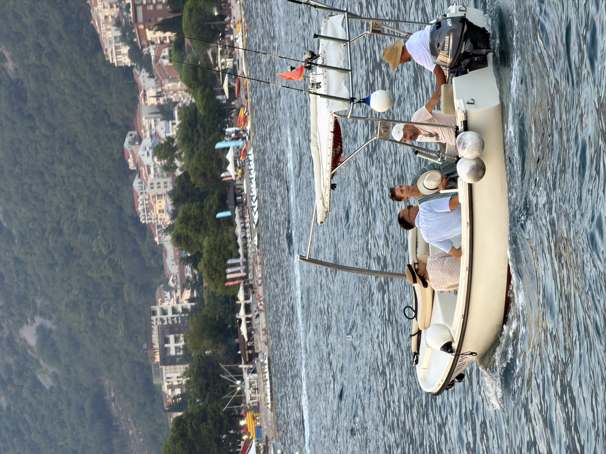 In Budva, Montenegro, a white motorboat with four people on board navigates choppy blue water. The boat has a Bimini top providing shade, with fishing rods mounted at the stern. The individuals appear to be enjoying a leisurely ride. In the background, a lush green hillside overlooks a coastline dotted with buildings, suggesting an urban coastal environment. Further out, a beach area with lounge chairs and umbrellas is visible, along with people enjoying various water activities. The sky is overcast, and the overall lighting suggests it is late afternoon or early evening.