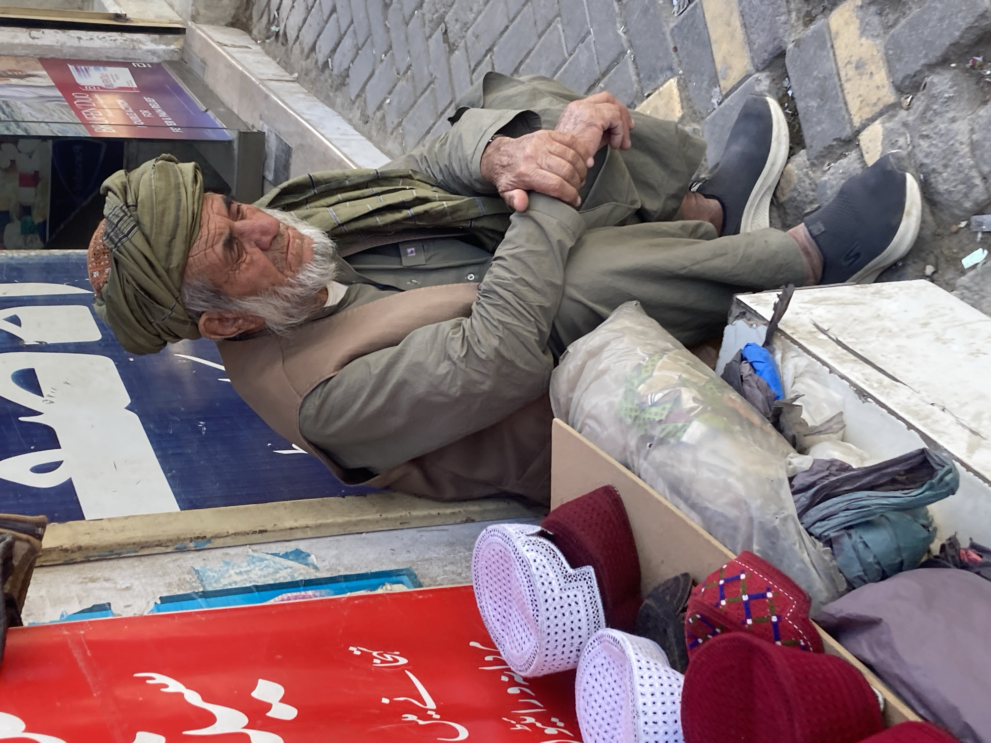 An elderly man with a turban, grey beard, and weathered hands is lying on the ground next to a red surface bearing Arabic script and a collection of hats. He wears a buttoned shirt, vest, and is shod in slip-on sneakers. The setting appears to be urban, potentially in Quetta City Tehsil, Pakistan, given the subject's dress and context of the scene.