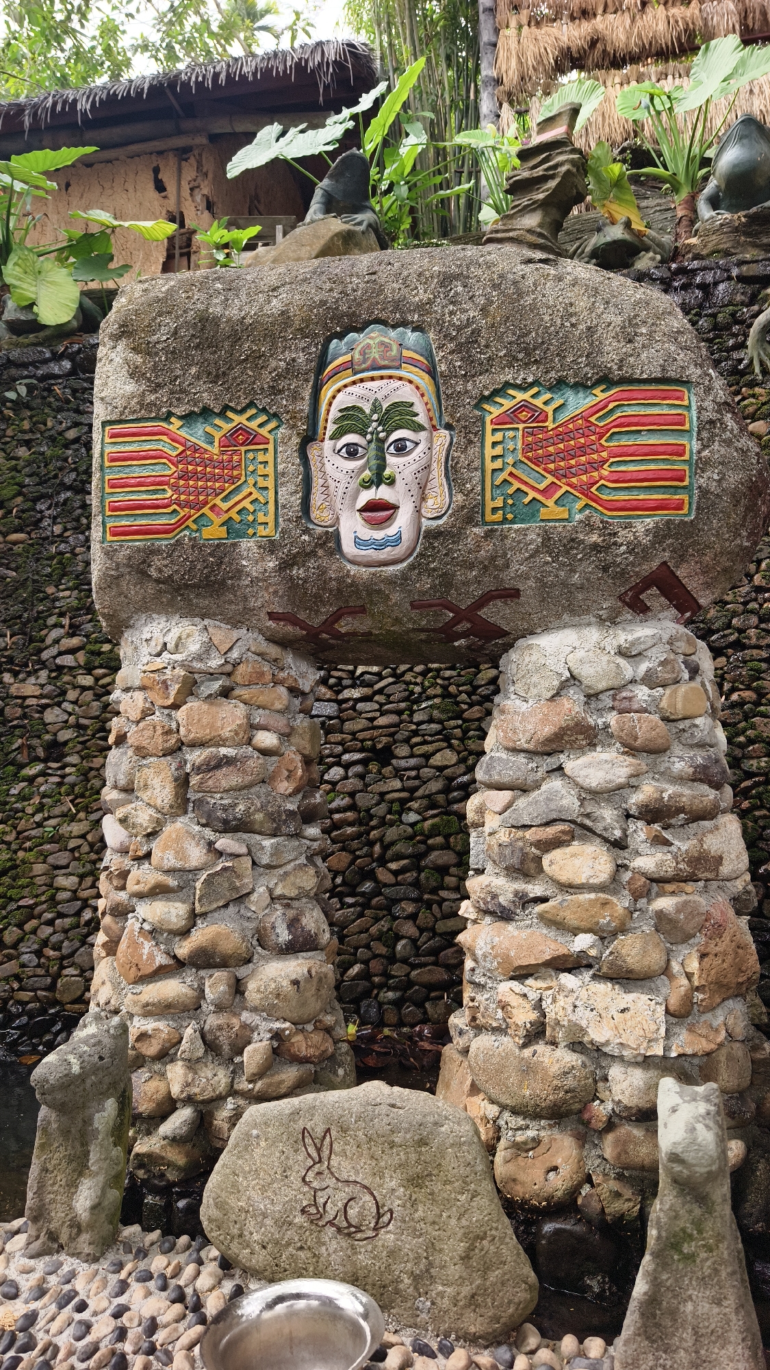 An outdoor scene in Jiyang District, China, depicts a decorative stone archway or monument integrated with natural elements. The central feature is a large, grey horizontal stone lintel supported by two substantial pillars constructed from irregularly shaped, cemented natural stones. The lintel displays three deeply recessed, vibrantly painted carvings: a stylized human face or mask in the center with green, white, red, black, and blue details, flanked by two symmetrical geometric motifs resembling stylized birds or fish, colored primarily in yellow, red, and green. Dark reddish-brown angular patterns are painted on the underside of the lintel between the pillars.
Behind the stone structure, a vertical wall is composed of numerous small, dark, smooth river stones, some exhibiting green moss, indicating the presence of moisture or running water that appears to flow downwards. Lush green vegetation, including large-leafed plants and tall bamboo stalks, densely populates the upper background. Portions of a building with a thatched roof are visible through the foliage. Several dark, rough-hewn stone figures, some appearing as abstract forms and others resembling frogs, are positioned on elevated ledges amidst the greenery.
The immediate foreground consists of a ground surface covered in small, smooth pebbles. Resting on these pebbles is a lighter-colored stone with an etched outline of a rabbit. Adjacent to it, a reflective, oval-shaped metallic basin is visible. Water is present at the base of the stone pillars, collecting in a dark pool. No human figures are present in the scene.