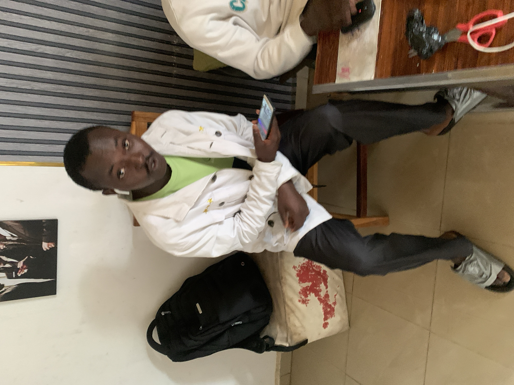 The image captures an indoor scene in Jos, Nigeria, featuring two individuals. The main subject, a young man, is seated on a wooden chair, looking directly at the camera with a neutral expression. He has dark skin and short dark hair, and is wearing a white collared jacket over a lime green t-shirt, black trousers, and grey slide sandals. White earbuds are visible in his ears, and he holds a smartphone with a lit screen in his right hand.
To his left, on the floor, rests a black backpack and a light-colored fabric bag or cushion with a prominent red, abstract pattern. The wall behind him features horizontal dark and light stripes, while an adjacent light-colored wall displays a framed, dark-toned picture.
Partially visible in the upper right is another person, seated at a wooden table next to the main subject. This individual is wearing a white long-sleeved top adorned with a light blue design. The wooden table holds various items, including a black computer mouse, red scissors, a black object wrapped in tape, and a white cable.
The scene portrays a casual, relaxed atmosphere, possibly during a break or a social gathering. The lighting suggests an indoor setting, with no visible text or explicit emotional cues from the subjects.
