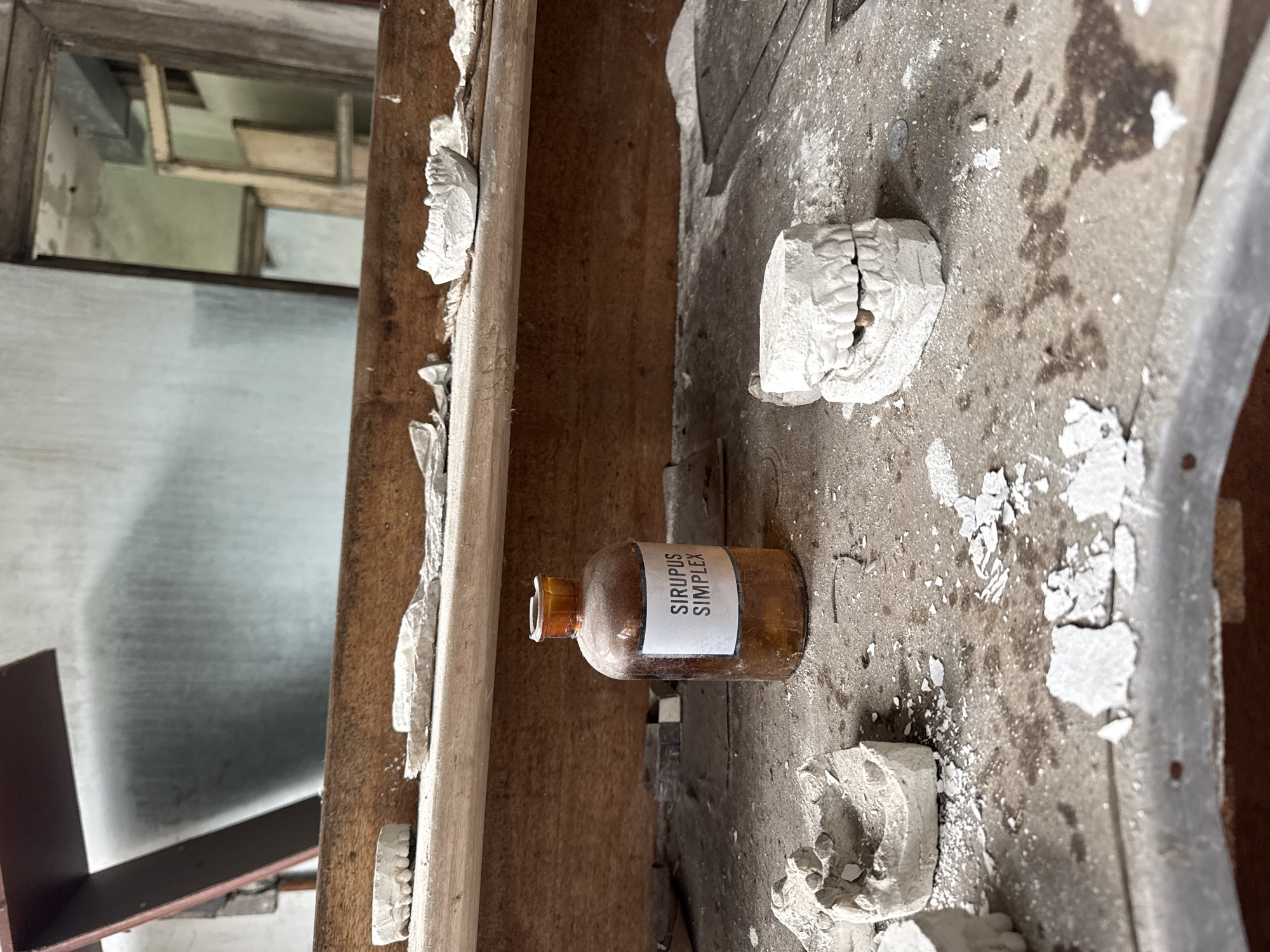 In a dilapidated interior, likely within the city of Prypiat, Ukraine, an amber-colored glass bottle labeled "Sirupus Simplex" rests on a dusty, deteriorating surface. The surface is covered in remnants of white material. Scattered around the bottle are fragments and shapes resembling dental molds. The background reveals a partially obscured window with a decaying frame. The overall environment is one of decay and abandonment.