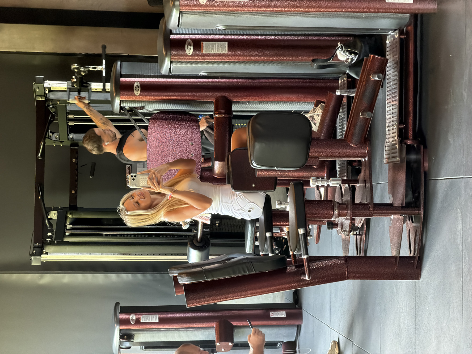 The image captures a gym scene in North Kuta, Indonesia. In the foreground, a woman with blonde hair is taking a mirror selfie with her phone, wearing a white top and skirt. She is positioned near a weight machine with red and metallic accents. Behind her, a man with short hair and a black tank top is using another weight machine, reaching for a handle. The gym environment is characterized by various exercise machines and grey tiled flooring.