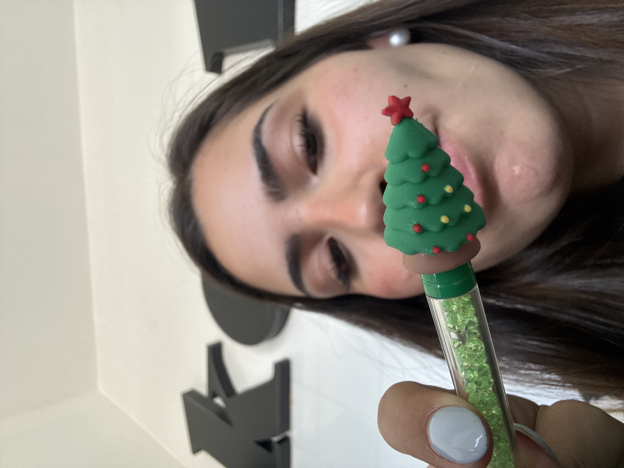 The image shows a woman holding a pen with a Christmas tree topper. The topper is green with red and yellow ornaments and a red star on top. The pen is clear and filled with green beads. The woman's face is visible in the background. The location is not discernible from the image.