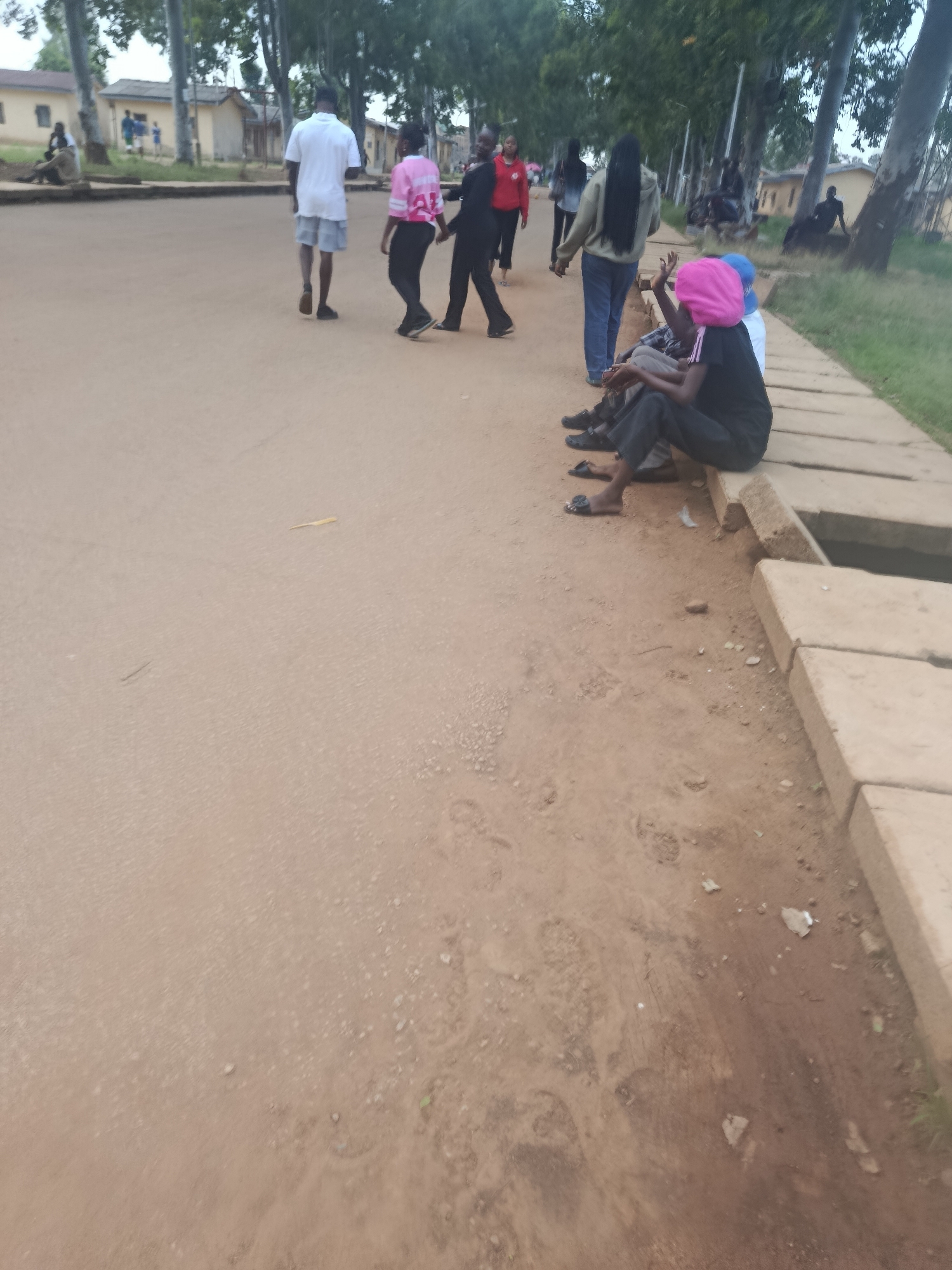 The image captures a scene on a dirt road in front of buildings, possibly in Rafin Par, Nigeria. Several people are present. In the foreground, to the right, a group of individuals is seated on concrete blocks bordering a drainage ditch. One person wears a bright pink head covering. Further up the road, a man in a white t-shirt and grey shorts walks away from the camera. To his left, a person in a pink and white top and black pants walks in the same direction. Other figures are visible in the background, including individuals walking and others seated near the buildings. The environment appears to be outdoors, with trees lining the left side of the road and the buildings on the far left. The ground is predominantly reddish-brown dirt, with some scattered debris. The lighting suggests it may be daytime, possibly overcast.