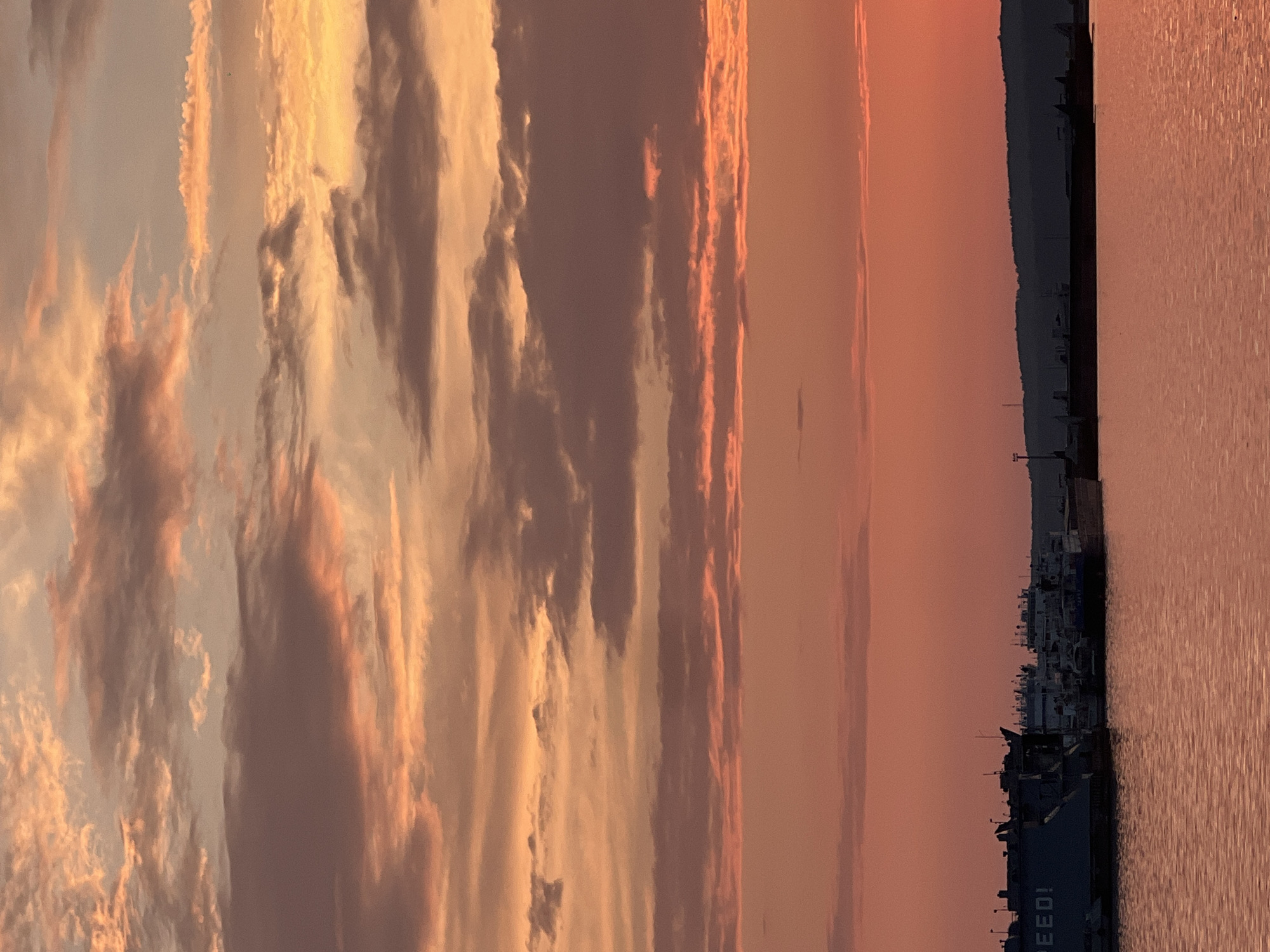 This is a photograph taken at sunset in the city of Chaikovsky, Russia, overlooking the Kama River. The sky is filled with dramatic clouds painted in hues of orange, pink, and purple, indicating the sun is setting. The water of the river reflects the colors of the sky. In the foreground, several docked ships are visible along the riverbank. One large vessel on the left has the partial word "SPEED!" printed on its side. The silhouette of a distant hilly landscape can be seen on the horizon. The scene is tranquil and captures the natural beauty of a sunset over a port.