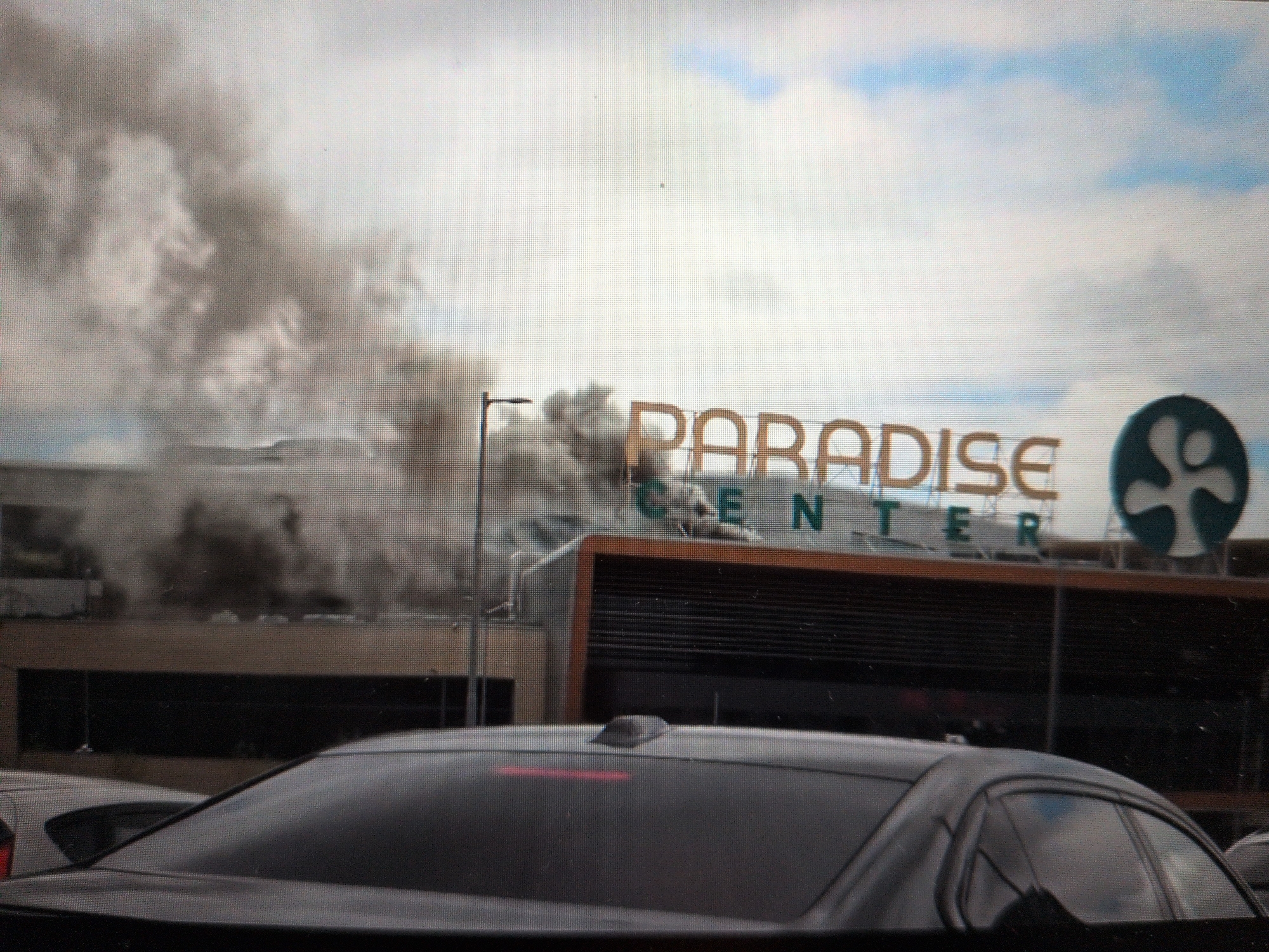 The image shows the Paradise Center shopping mall with smoke billowing from the rooftop. The mall's sign is visible, along with its logo. In the foreground, the rear of a dark-colored car is visible. The sky is cloudy. The location is likely in an urban or suburban area due to the presence of the shopping mall and cars.