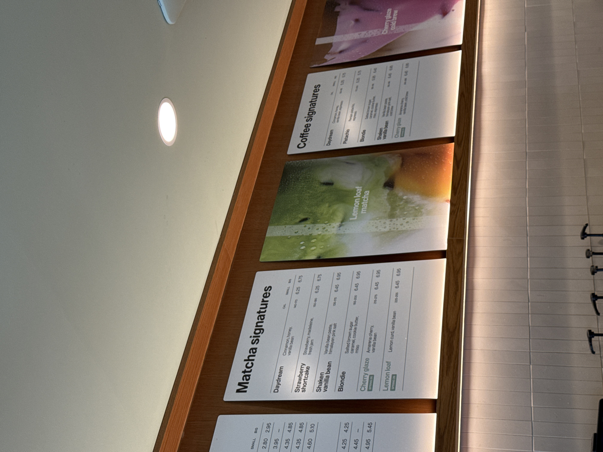 The image shows a menu board, likely inside a cafe or similar establishment. The board is divided into sections for "Matcha Signatures" and "Coffee Signatures." The Matcha section lists various drinks like "Daydream," "Strawberry shortcake," "Shaken vanilla bean," "Blondie," "Cherry glaze," and "Lemon loaf Matcha." Each drink is listed with a brief description of its ingredients and calorie count, along with small and big size pricing. The Coffee section lists drinks like "Daydream," "Pistachio," "Blondie," "Shaken vanilla bean," and "Cherry glaze cold brew." The board is mounted on a wooden frame against a light-colored tiled wall. The lighting is bright and appears to be from recessed ceiling lights. The image does not provide sufficient information to determine the location of the establishment in Boston.