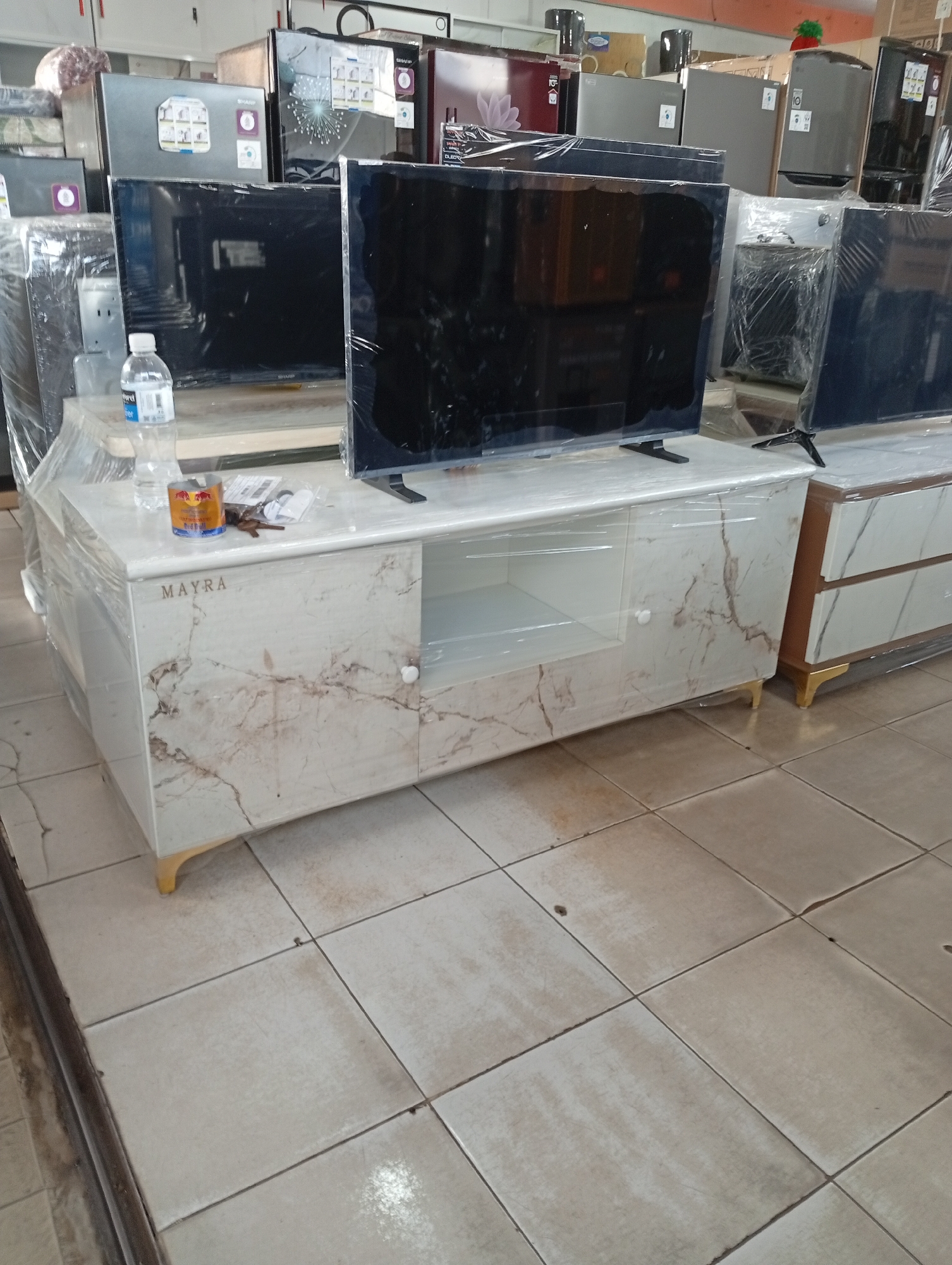A store in Batam, Indonesia, displays televisions and refrigerators for sale. The televisions are black, flat-screen models, some wrapped in plastic. They are placed on white marble-patterned furniture with gold legs. In the background, several refrigerators, in colors such as silver, black, and red, are also visible. A bottle of water and a red bull can are on the furniture. The floor is tiled with off-white squares. The overall scene suggests a retail environment focused on home appliances.
