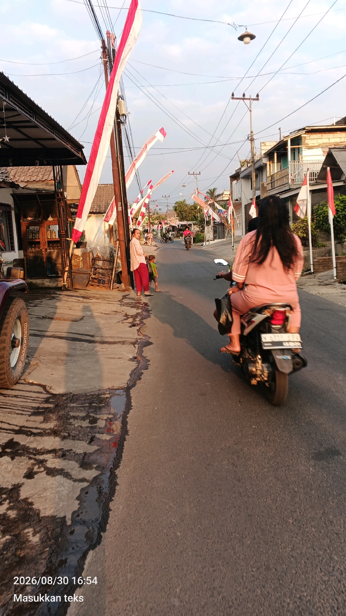 The image depicts a street scene in Jeru, Indonesia, adorned with Indonesian flags, suggesting a celebratory or patriotic occasion. In the foreground, a portion of a maroon vehicle's tire is visible on the left. The street is paved, with a noticeable patch of water and debris on the left side of the asphalt. A woman in a pink outfit is riding a motorcycle in the right lane, with her back to the camera. Further down the street, other individuals and motorcycles are present, with more flags and banners lining the road. Buildings with traditional Indonesian architecture are visible on both sides of the street. The sky is clear with hints of clouds, and the setting sun casts long shadows. A timestamp "2026/08/30 16:54" and the text "Masukkan teks" are superimposed on the bottom left corner of the image.