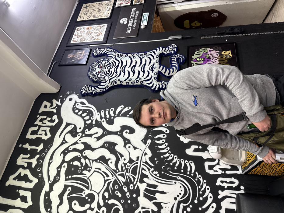 The image captures a man standing within an artistically decorated indoor space, likely a tattoo studio or an art gallery. The walls are painted black, serving as a dark backdrop for various artworks and decorations.
A man, appearing to be in his twenties or early thirties, stands slightly to the right of the center, looking directly at the camera. He has short, dark brown hair and a neutral expression. He is wearing a light grey hooded sweatshirt featuring a small blue fish logo embroidered on the left side of his chest. A black strap from a bag crosses his body, leading to a green utility-style messenger bag worn at his hip. A green bottle, possibly a drink, is partially visible in a side pocket of his bag.
The black wall behind him is adorned with several distinct features. On the lower left side, a large, intricate mural in white paint depicts stylized, flowing lines and shapes that resemble abstract or artistic tiger patterns. The words "OLD TIGER" are partially visible in a bold, graffiti-like font within this mural on the far left.
Above the man's head and to his left, a white and blue striped tiger-shaped rug or plush wall hanging is prominently displayed on the black wall. The tiger has red eyes.
Further up the wall, several framed artworks are hung. One rectangular sign clearly displays the text "OLD TIGER TATTOO x MOSCOW OFFENDER". Other framed pieces appear to be tattoo flash sheets or art prints featuring various intricate designs. To the right of the tiger rug, another framed piece of art showcases abstract shapes and colors, including purple, green, and yellow, against a dark background.
In the upper right background, a doorway or opening leads to another section of the room. On the floor in this area, a dark mat shaped like the BAPE (A Bathing Ape) logo is visible. Above this opening, a small white sign with black text reads "ВЫХОД", which is Russian for "EXIT". The lighting suggests an indoor environment, likely illuminated by artificial lights. The overall aesthetic implies a creative and possibly urban-influenced setting.