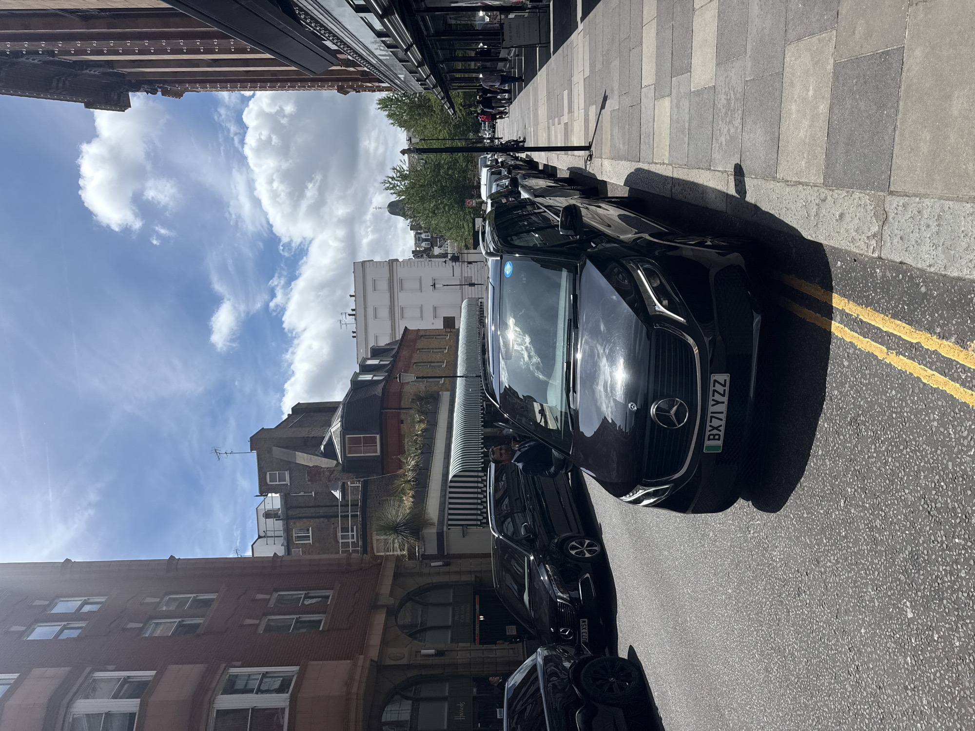 The image, oriented vertically, captures a street scene in London, United Kingdom. The foreground is dominated by the front of a black Mercedes-Benz car, its license plate clearly visible as "BX71 YZZ". Adjacent to the car, the road surface is marked with double yellow lines. Beyond the car, the sidewalk is paved with large, grey tiles.
Further down the street, a line of parked black cars stretches into the distance. People are visible walking on the sidewalk, some in groups, and one person is partially visible in the driver's seat of one of the parked cars. Behind the parked cars, the facades of buildings line the street. The architecture suggests a historic urban setting, with brickwork, arched doorways, and large windows. Some buildings have awnings or canopies over their entrances. In the background, trees with green foliage are visible, and further still, more buildings, including some with distinctive domed roofs, can be seen against a bright blue sky with scattered white clouds.
The time of day appears to be daytime, likely late morning or early afternoon, given the bright sunlight casting shadows. The weather seems pleasant, with a clear sky and sunshine. The overall impression is of a typical day in a busy urban environment. One building in the middle distance displays a sign that reads "Harrods," providing a specific location cue. Another sign, partially obscured, appears to be for a hotel or establishment. The overall mood is neutral, depicting a common urban landscape.