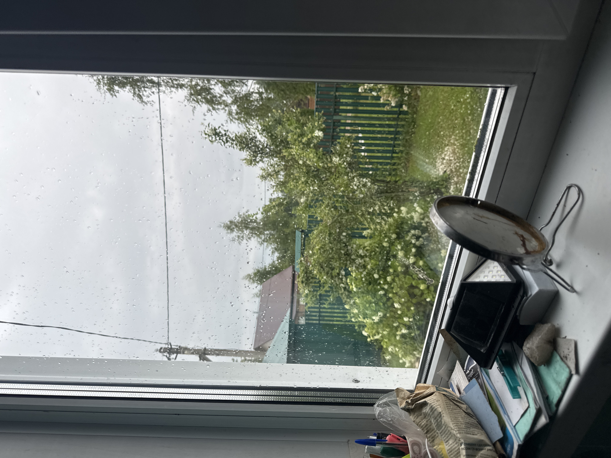 The image depicts a view from a window in Красное, Russia, on a rainy day. Raindrops are visible on the glass, obscuring the outdoor scene to some extent. Outside, a green wooden fence encloses a yard with lush green grass and flowering bushes. Beyond the fence, a house with a brown roof and a green-roofed structure are partially visible, surrounded by trees. On the windowsill, various objects are scattered, including pens in a holder, crumpled paper, a small black electronic device with a solar panel, a clothespin, and a small rock. A round mirror with a metal stand is also present on the sill. The overall impression is one of a quiet, domestic scene observed during inclement weather.