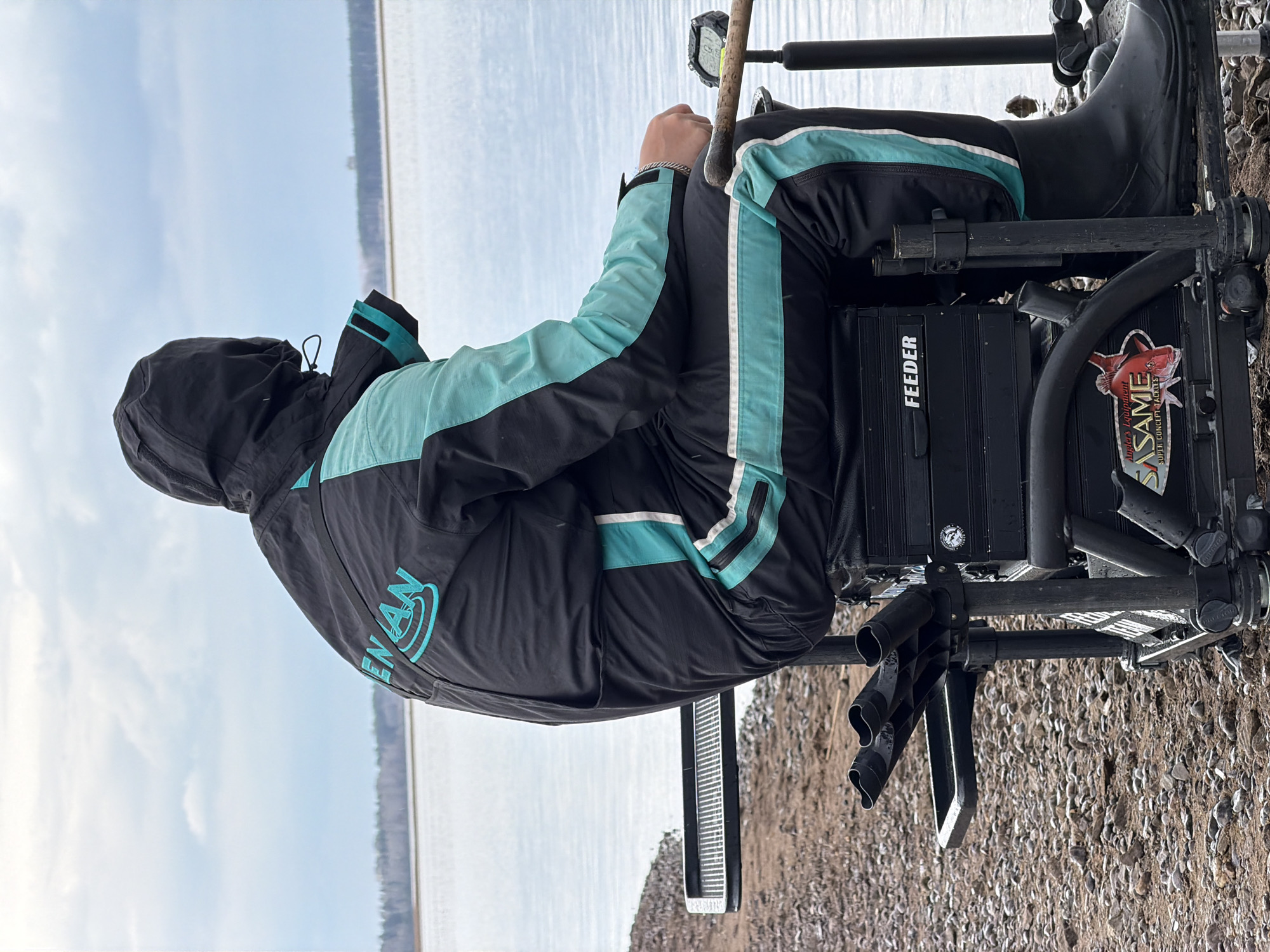 A person in a black and light blue jacket and black boots sits in a black metal fishing chair on a pebble-covered shore. The person is facing away from the camera and toward a large body of water under a cloudy sky. The chair has the word "FEEDER" on the front of it and "SASAME" on the side. The location is Chaikovsky, Russia.