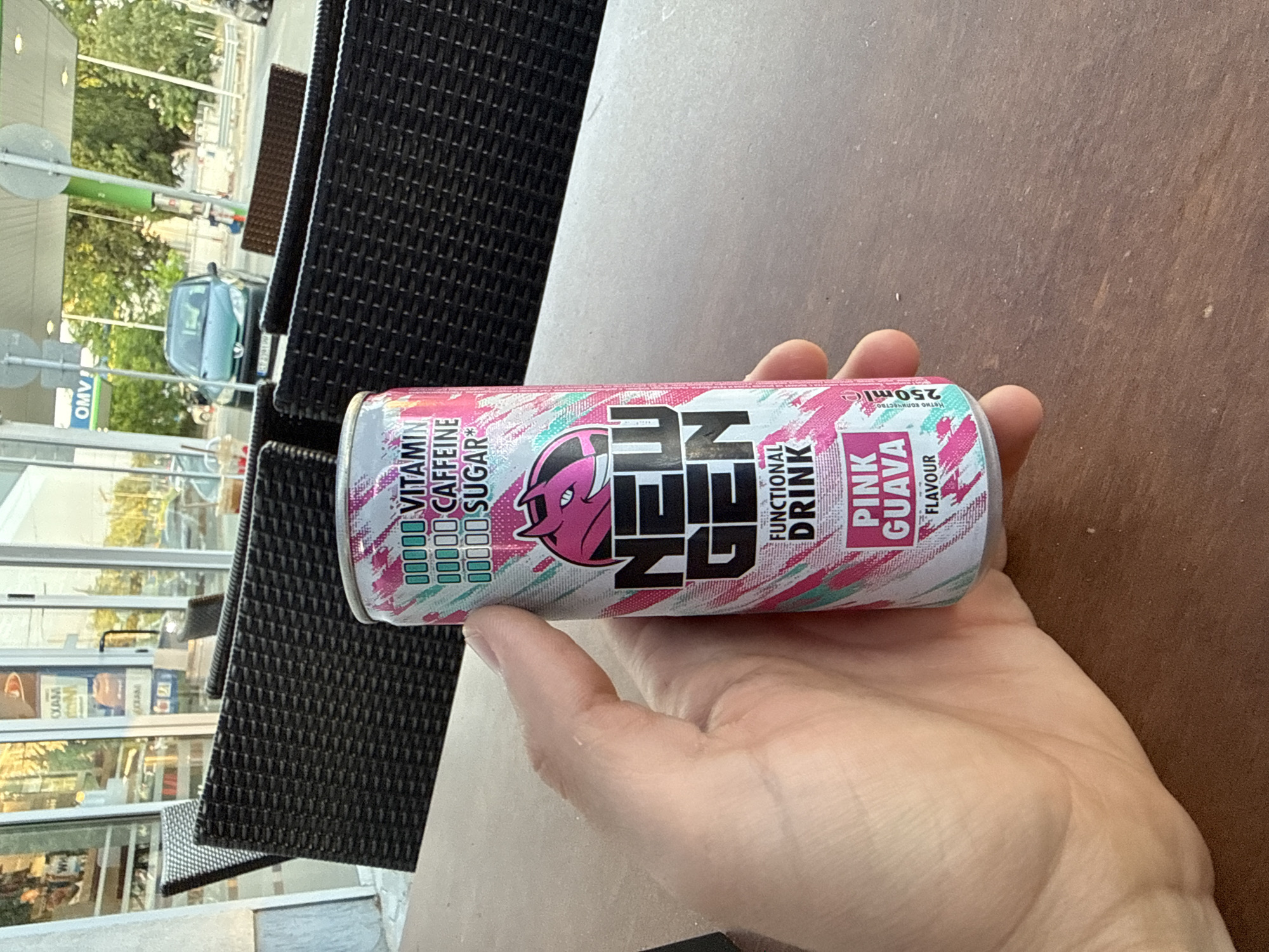 A right human hand, with visible skin creases and minimal fine hairs, holds a 250ml "NEW GEN" functional drink can against a light brown wooden table. The can features a pink, white, and teal abstract design, displaying "NEW GEN" in large black letters, "FUNCTIONAL DRINK," and "PINK GUAVA FLAVOUR." A stylized pink devil-like head logo with horns and sharp teeth is positioned above the brand name. Below the can's rim, square icons are present, indicating "VITAMIN," "CAFFEINE," and "SUGAR." The background depicts an outdoor seating area, typical of a cafe or petrol station convenience store in Stara Zagora, Bulgaria. Several dark-brown, woven-patterned outdoor chairs are arranged, partially obscuring a building with large glass windows on the left. To the right, an "OMV!" branded petrol station canopy with a green supporting pillar is visible. A dark green Peugeot car with a Bulgarian license plate "CD 2391 MM" is parked under the OMV canopy. Trees and additional vehicles are discernible further in the distance, under a bright, daylight sky.