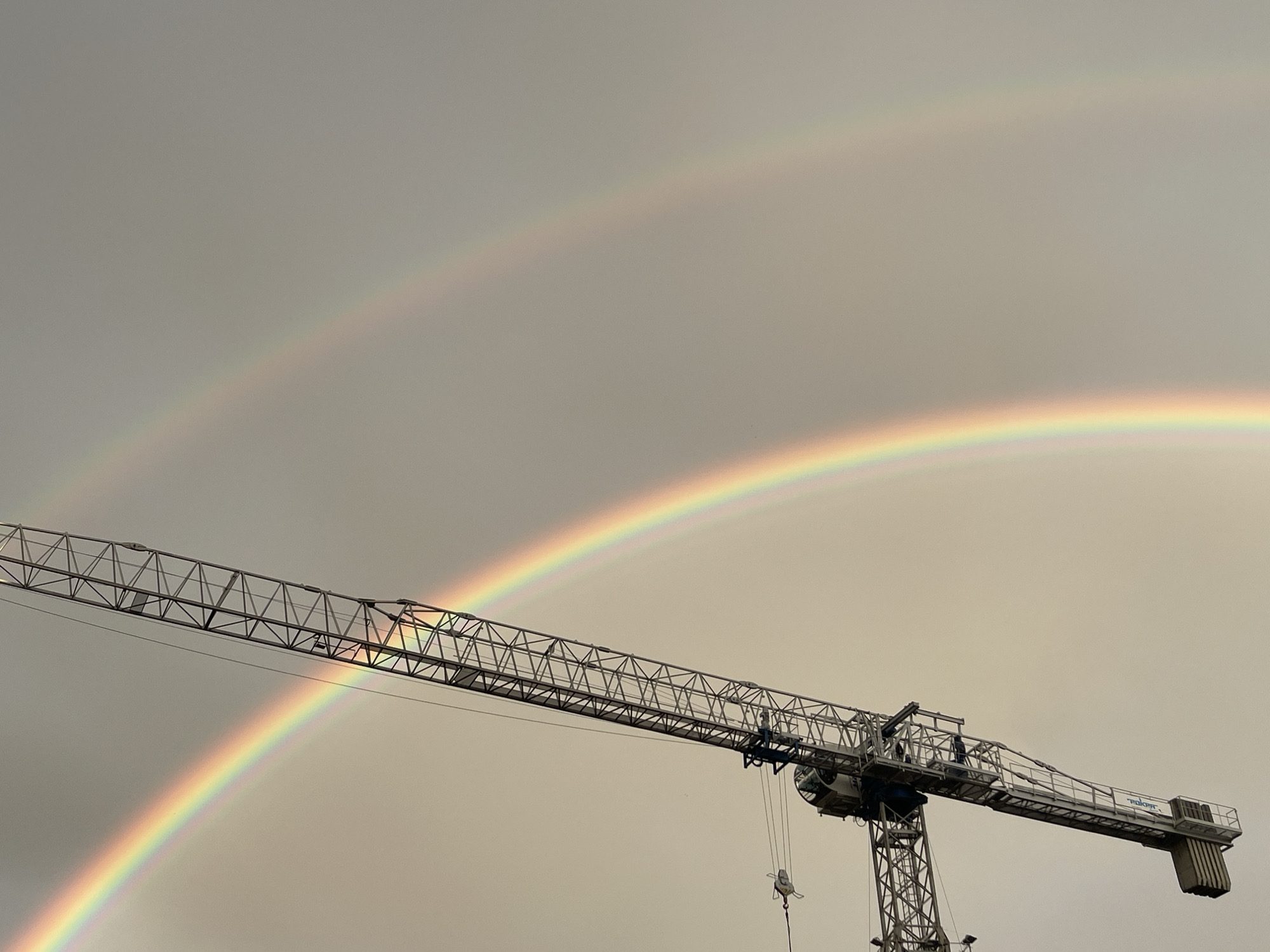 An outdoor, eye-level, wide shot captures a double rainbow against a hazy, overcast sky. The larger, lower rainbow is vibrant with distinct colors, while the smaller, upper rainbow is fainter. A large construction crane bisects the frame diagonally from the bottom left to the top right, its metal lattice structure silhouetted against the sky. The crane is positioned in the foreground, with its jib extending horizontally across the image. The overall impression is one of a construction site illuminated by a natural phenomenon.