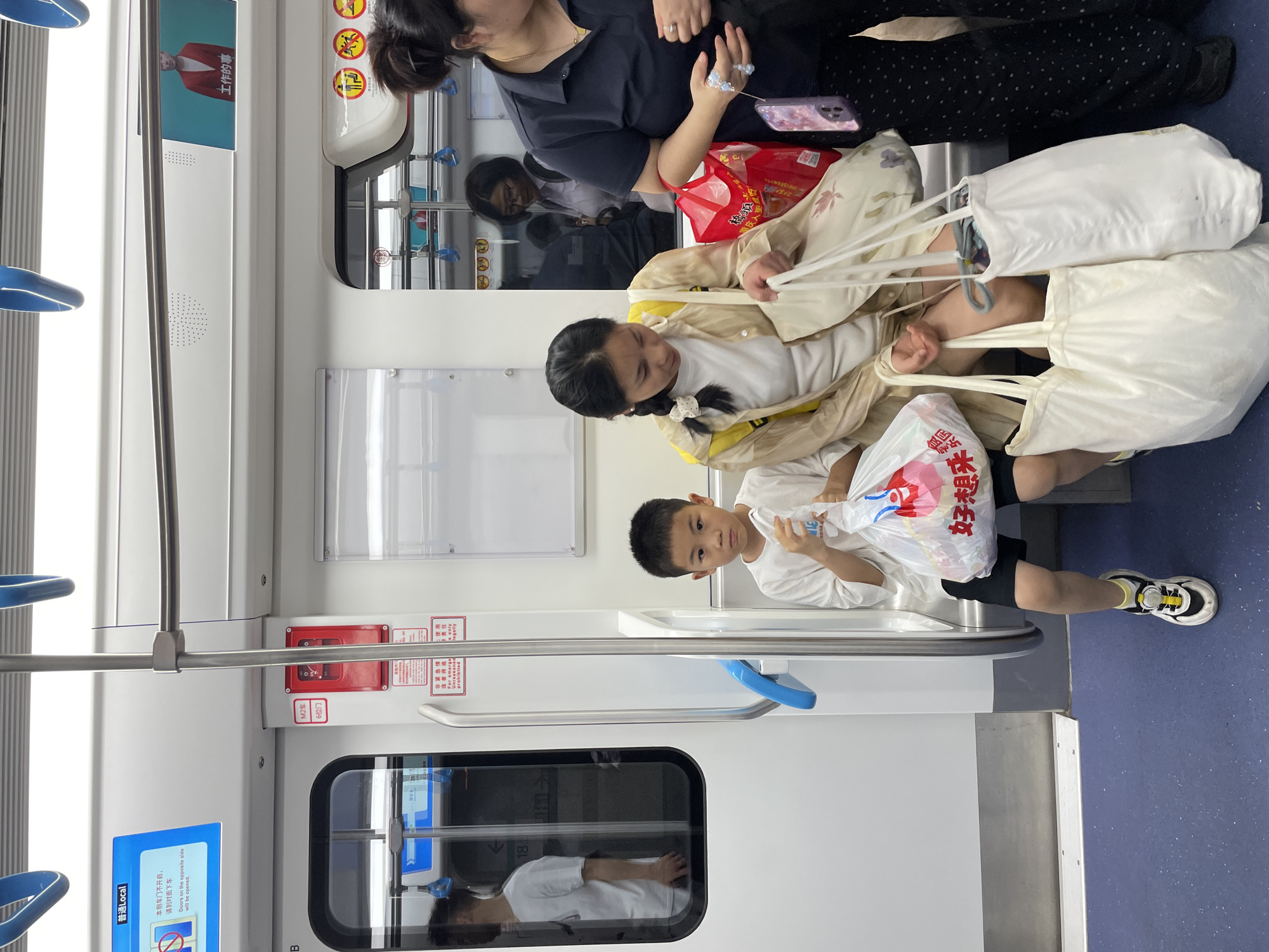 The image shows the interior of a subway car. Several people are present, including an adult woman, a teenage girl, and a young boy. The teenage girl is seated and holding several white tote bags. The young boy is standing next to her and holding a plastic bag with red and white writing. The adult woman is standing above them and holding a cell phone. The subway car has a window with a reflection of a person, and a blue panel with white text in Chinese. The environment is brightly lit and clean. The image likely originated in a city in China, based on the text.