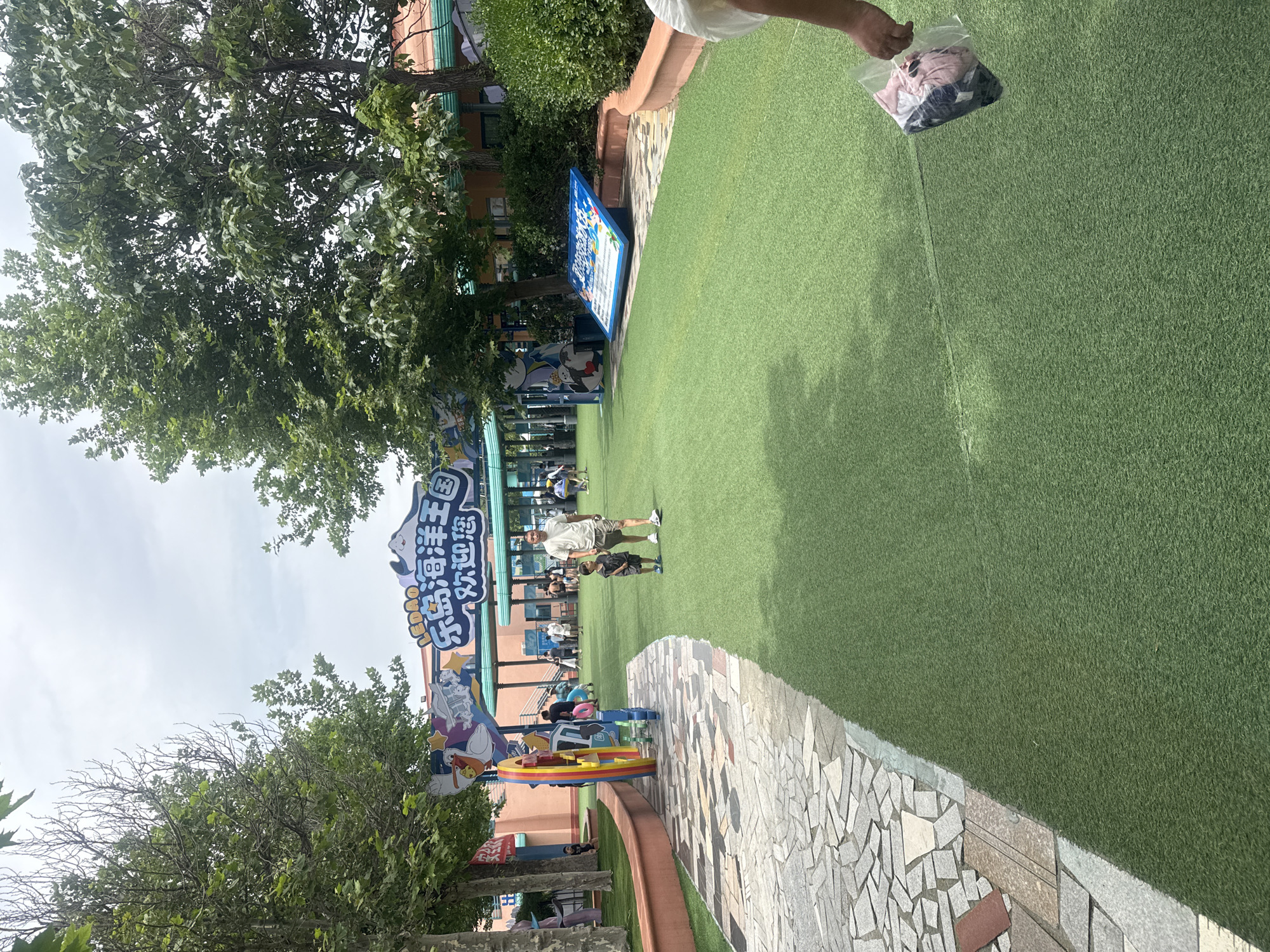 The image depicts an outdoor scene at an amusement park in Qinhuangdao, China. A man and a young boy are walking on artificial turf. In the background, there is an archway with Chinese characters and cartoon figures, likely serving as an entrance to the park. Several other people are visible in the distance, and trees and foliage surround the area. A blue information board is propped up on the grass. The overall atmosphere suggests a recreational setting on a cloudy day.